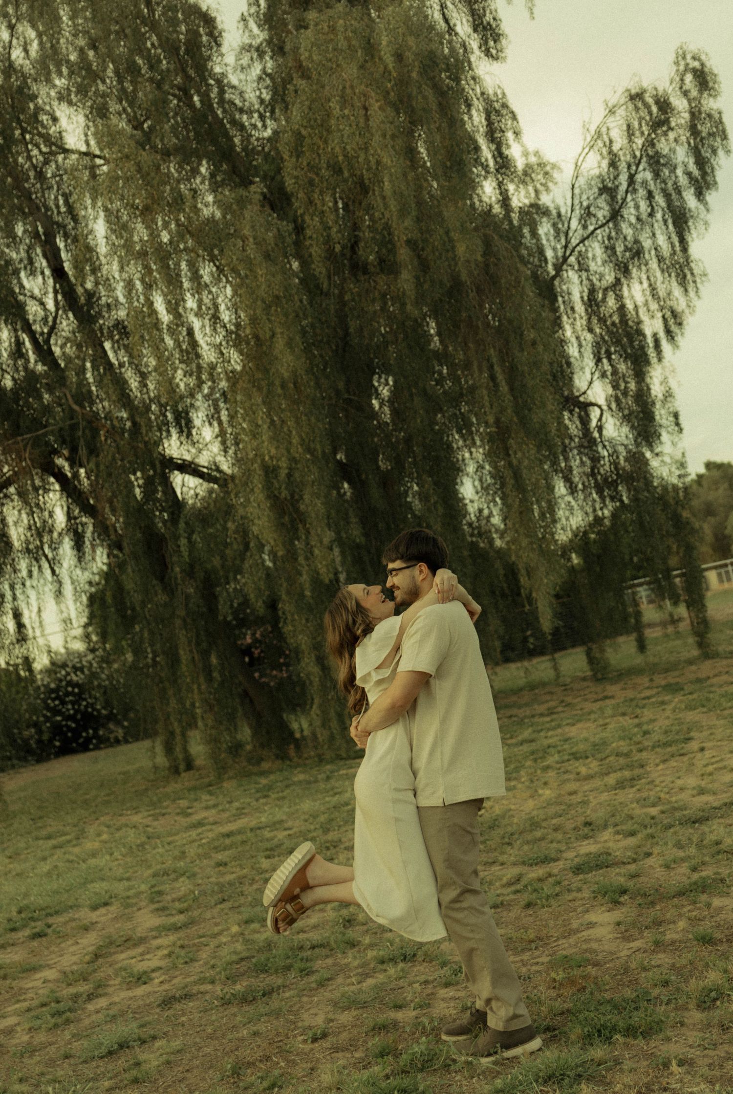 Man lifting woman in a joyful embrace under a weeping willow tree in a park.