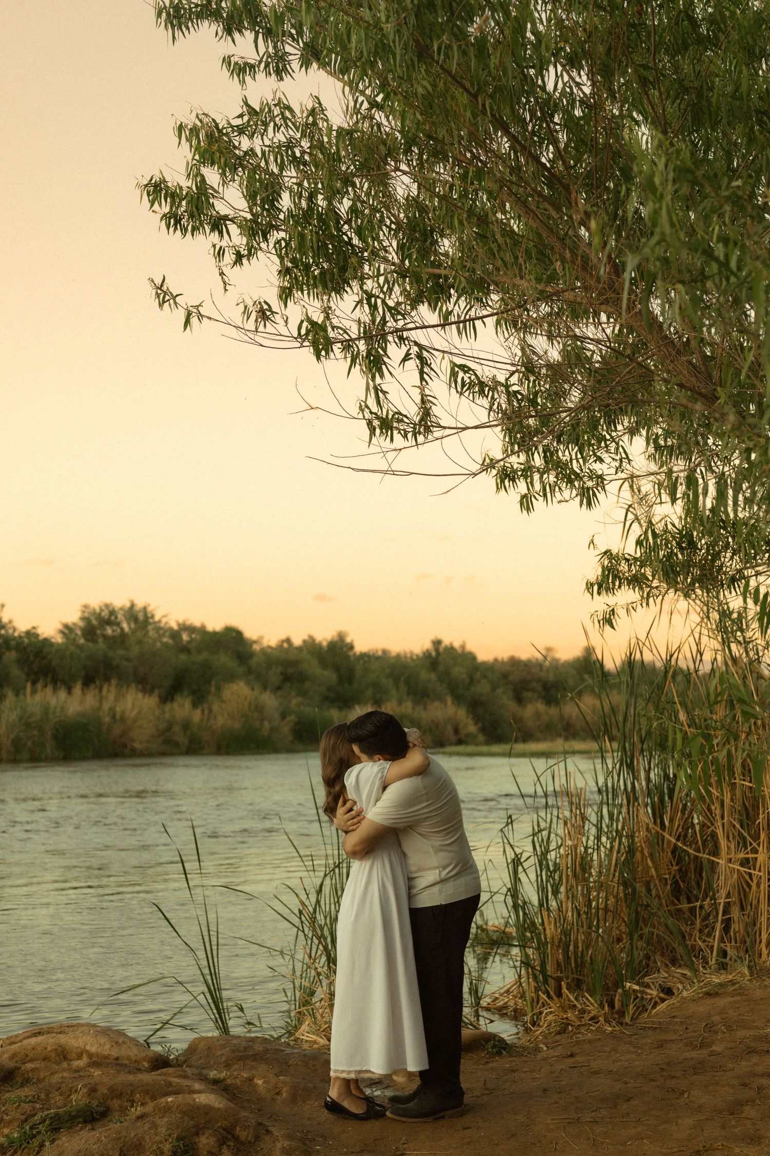 Couple sharing a tender embrace under a willow tree by a river at sunset.