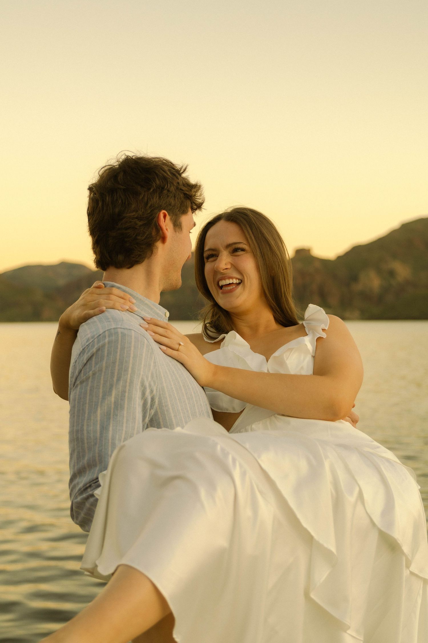 A couple in white clothing embrace and laugh together during sunset by the water.