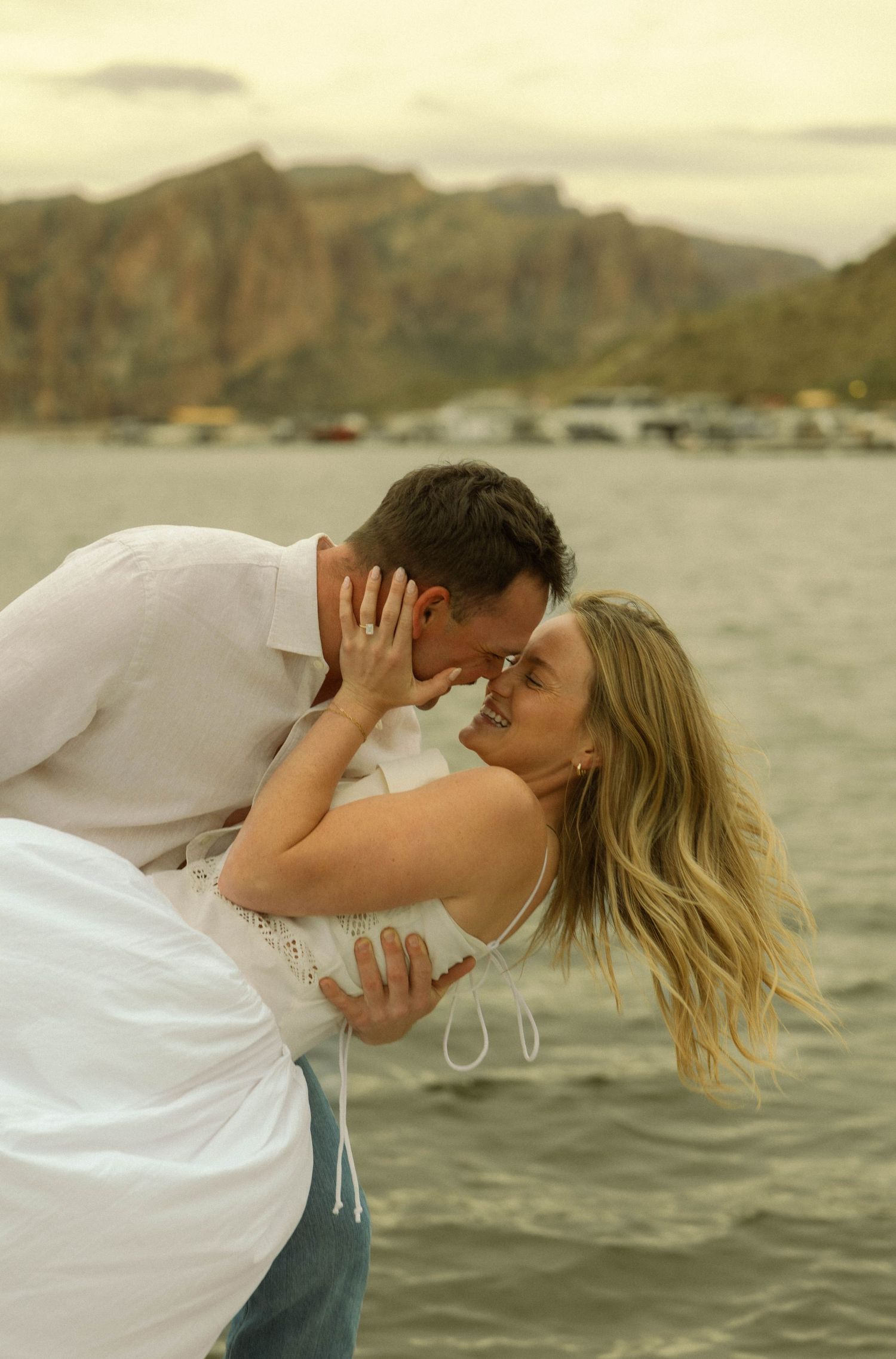 A romantic dip pose by a couple in white clothing with mountains and water in the background.