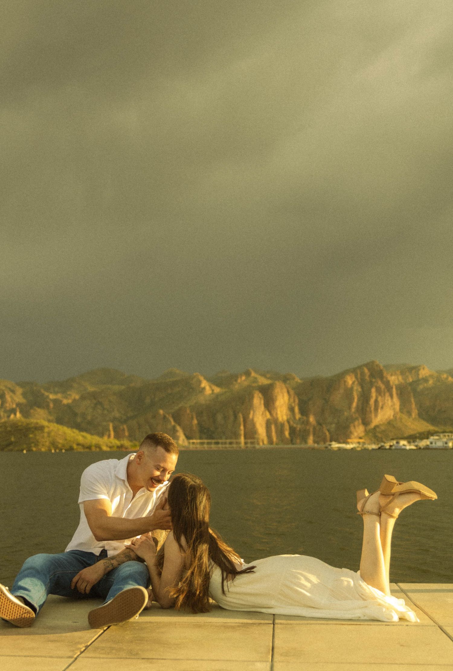 Couple sharing a romantic moment on a boat deck with dramatic coastal cliffs and mountains in the background.