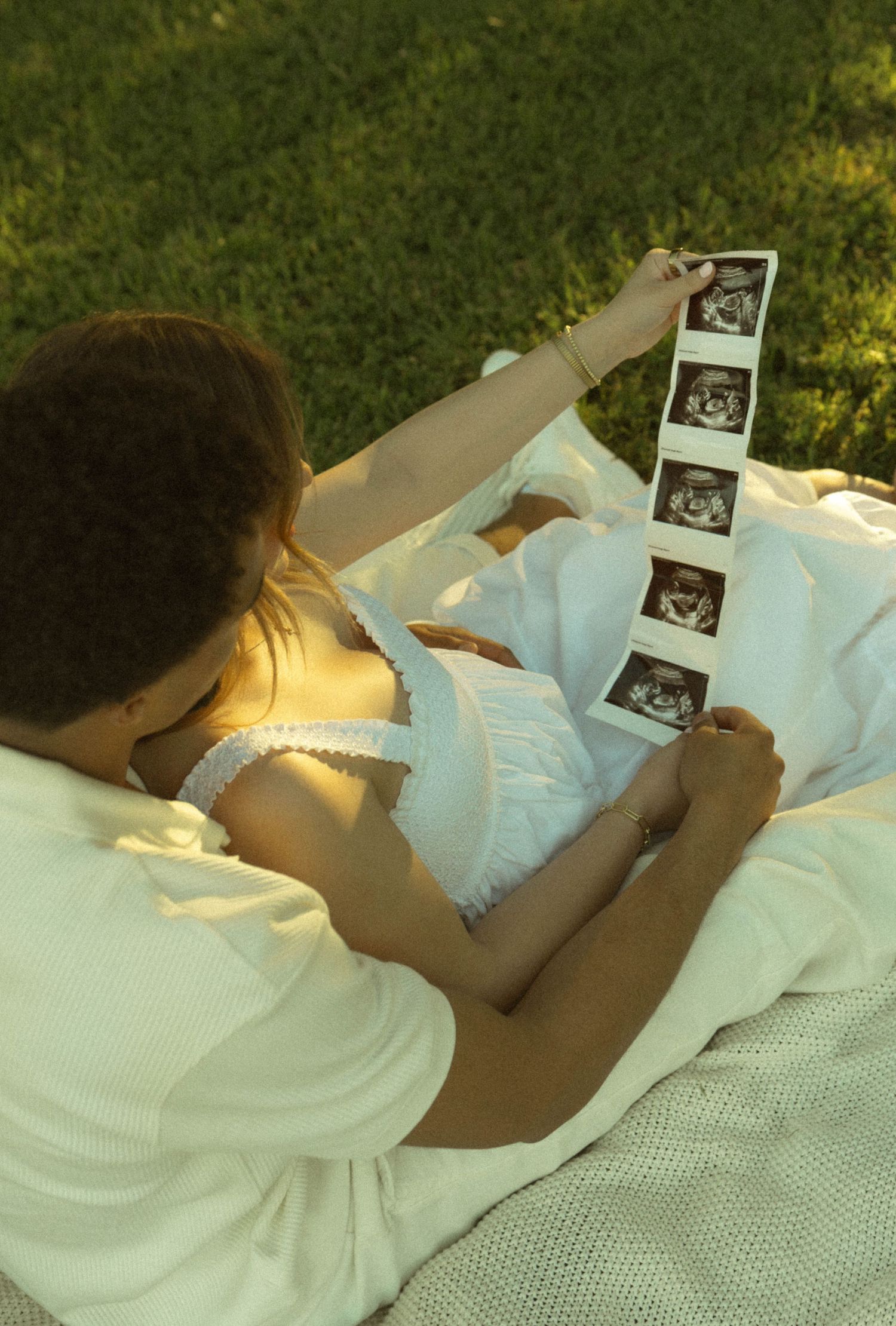 Couple lying on blanket in grass reviewing ultrasound photos together outdoors.