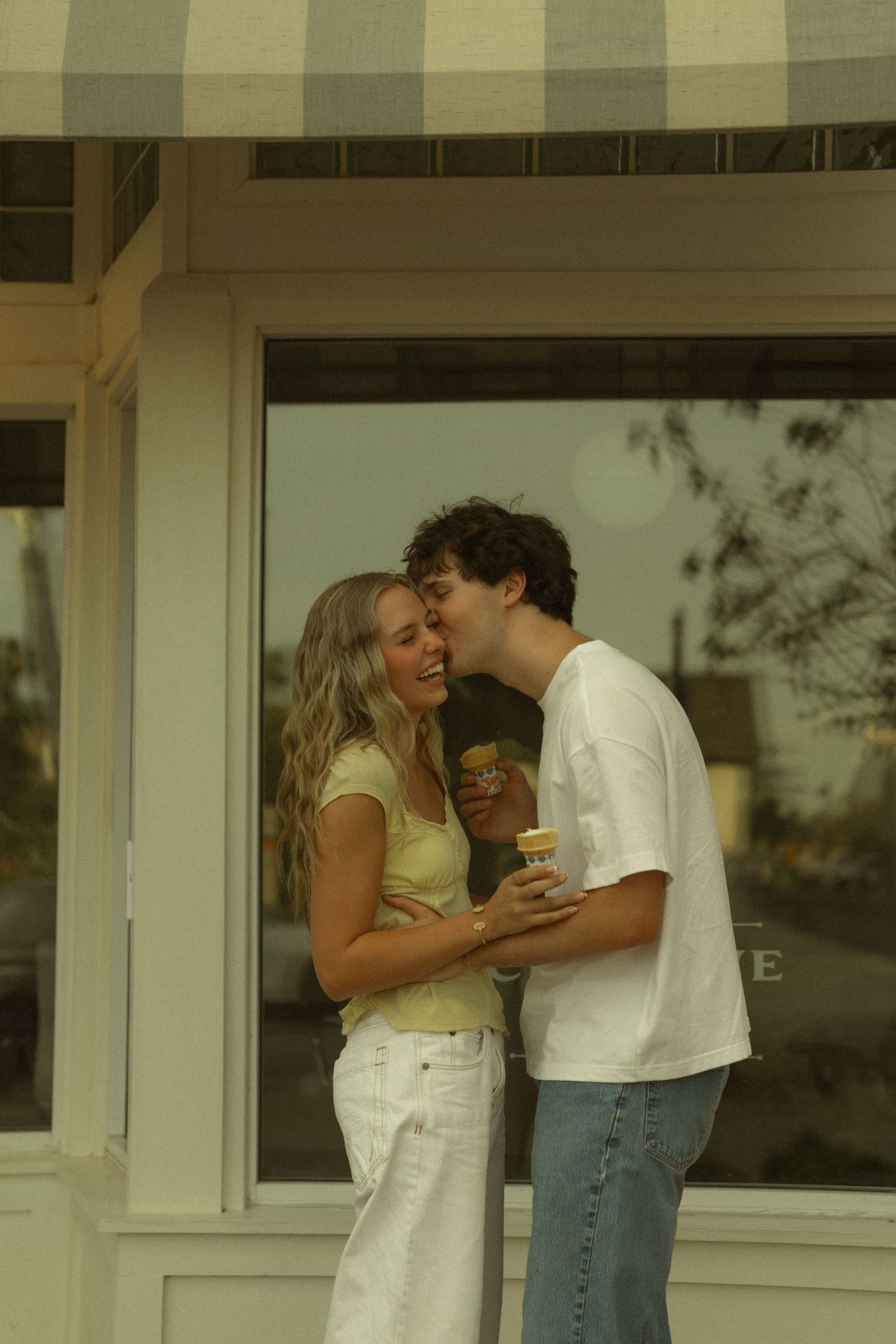 A happy young couple laughs and shares ice cream cones outside a charming striped awning shop.
