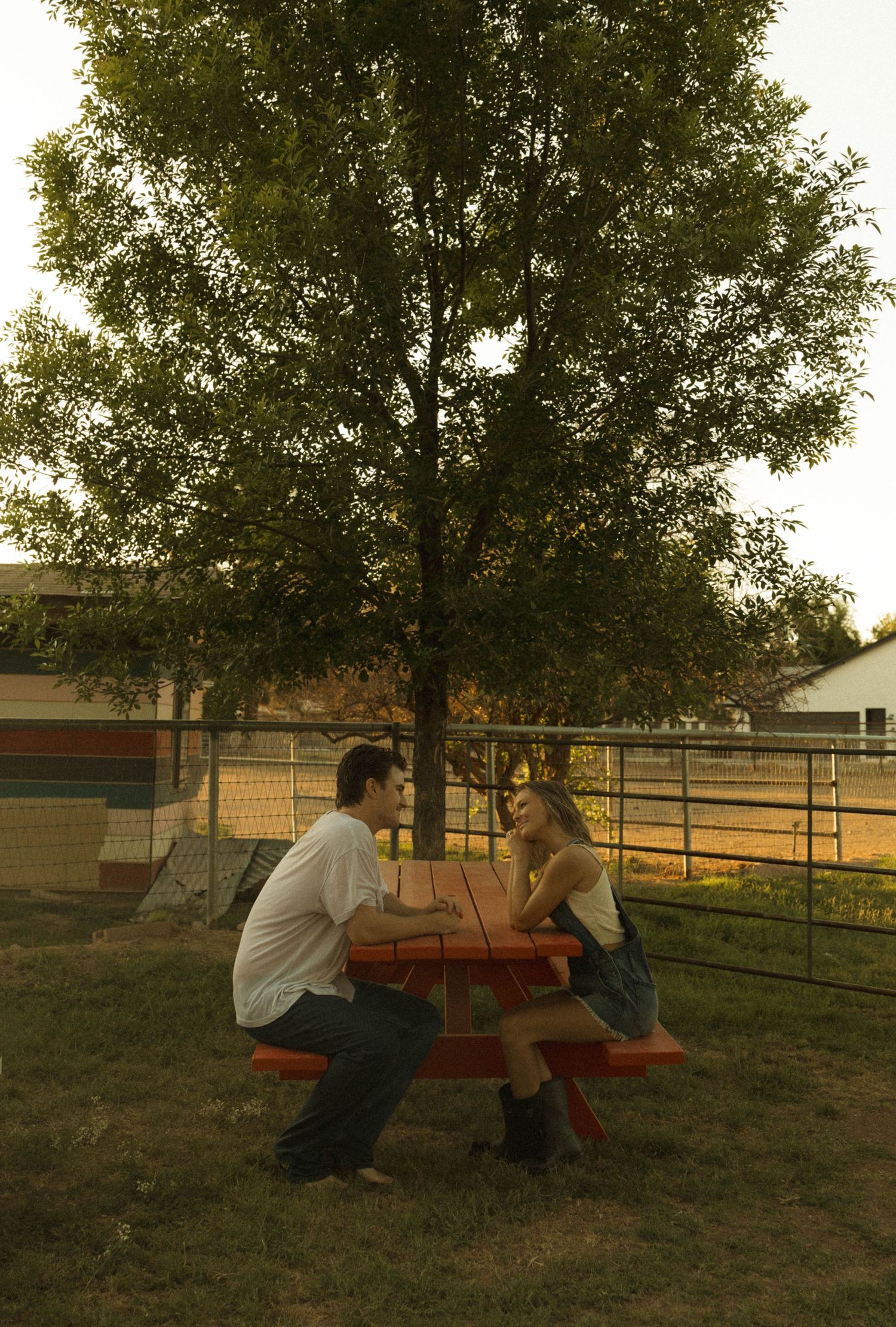 A young couple sits across from each other at a red picnic table under a large tree at dusk.