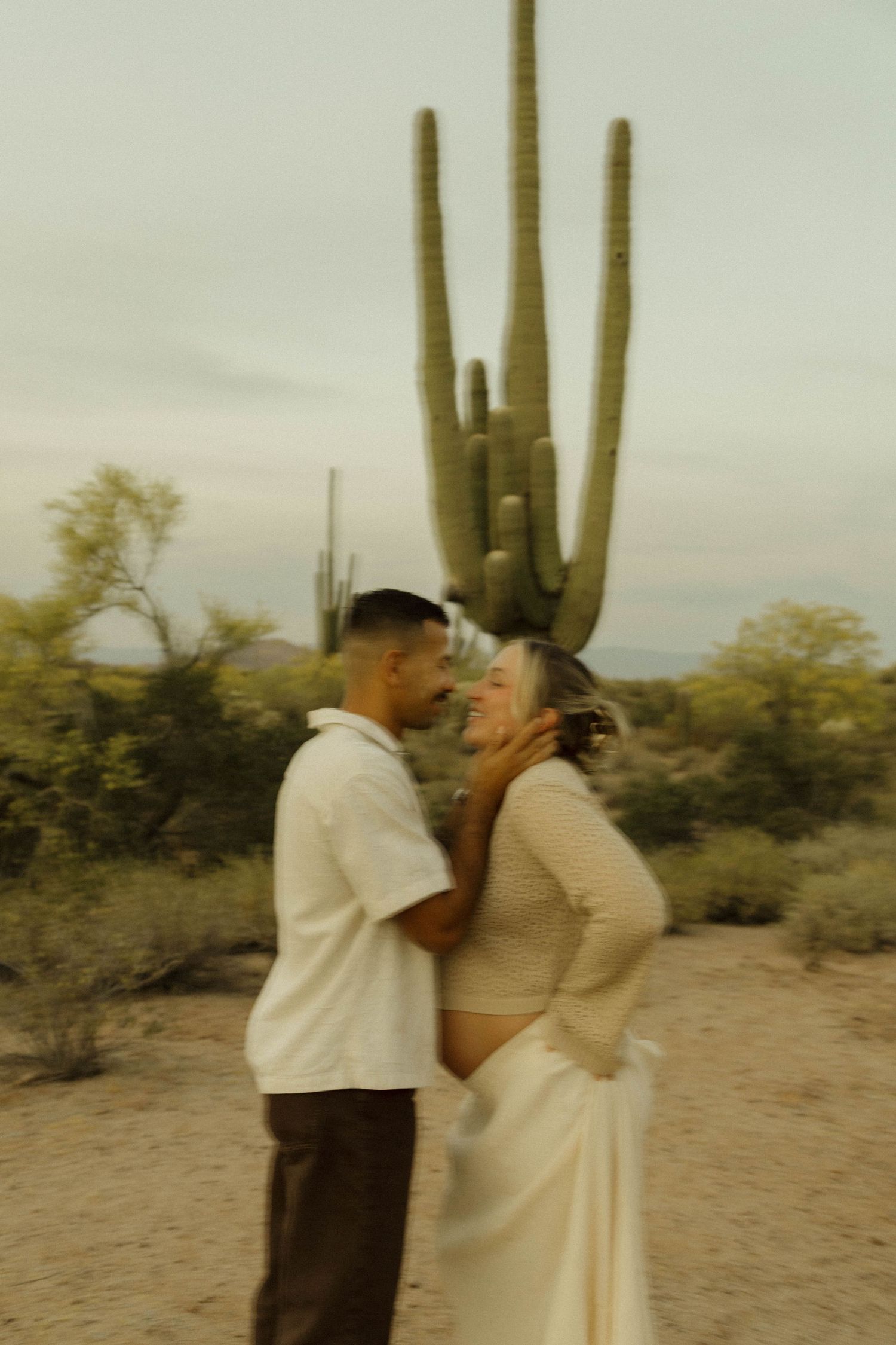 Couple sharing romantic moment in desert with saguaro cactus, woman in white skirt and lace top.