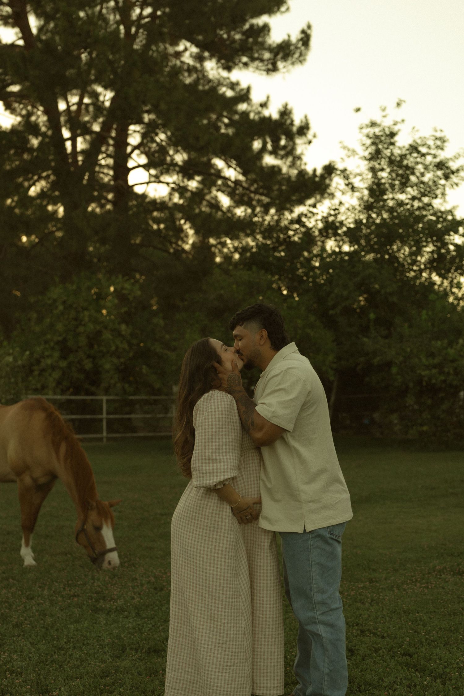 Couple sharing tender moment in field with grazing horse at sunset.