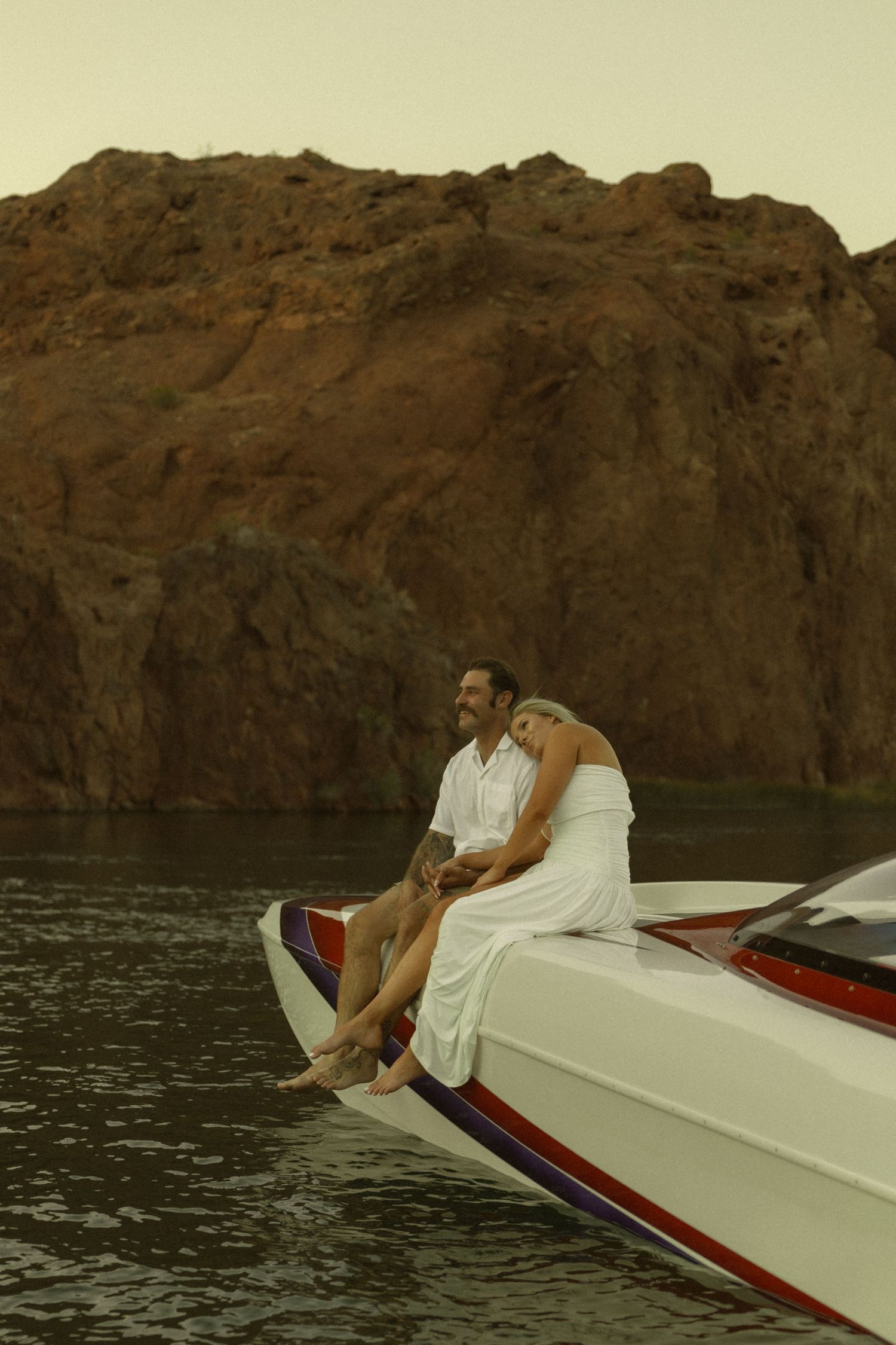 Couple relaxing together on speedboat bow with legs dangling over water near rugged rock formation.