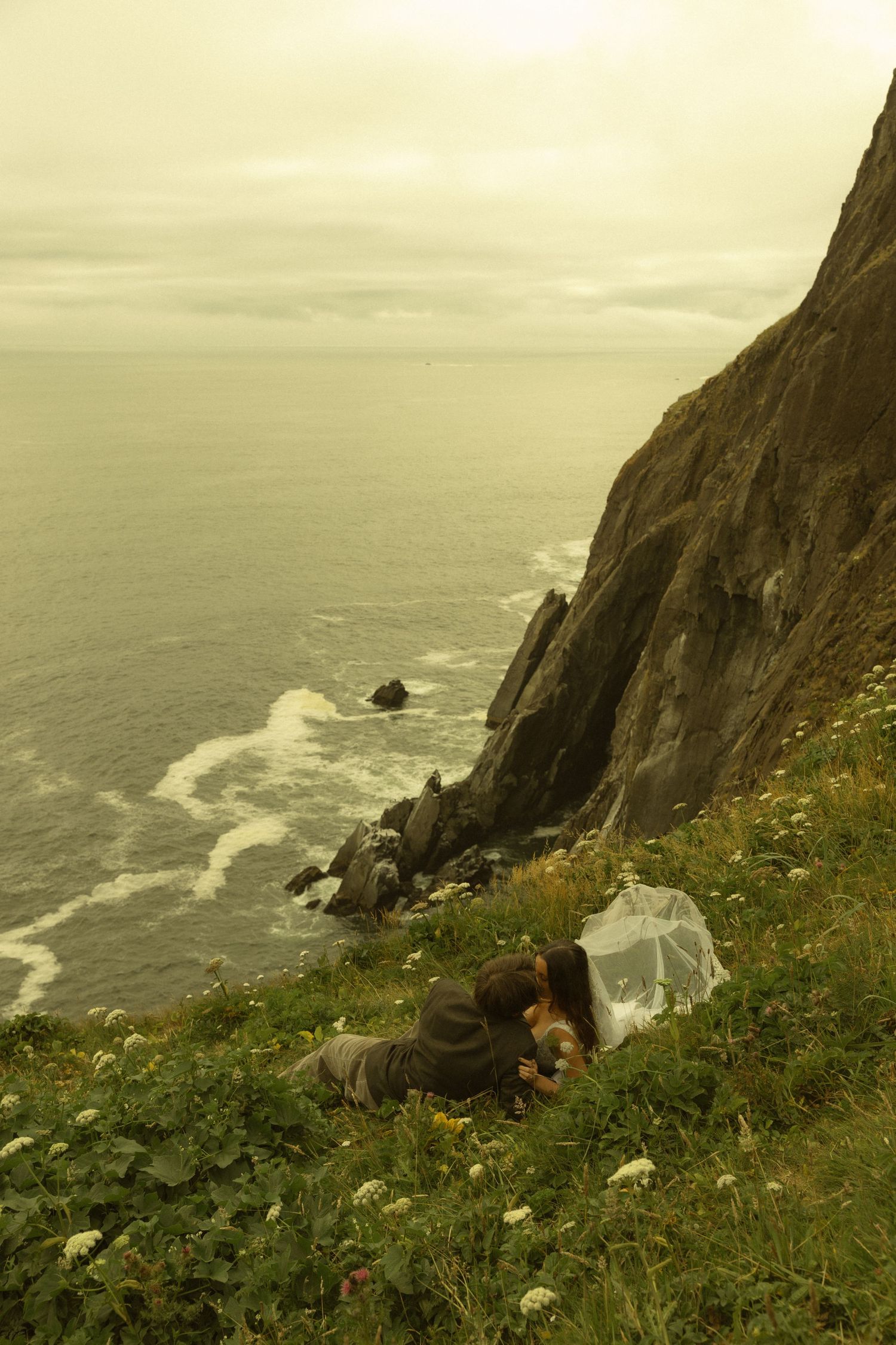 Couple sitting on a cliffside meadow overlooking rocky ocean coastline with waves crashing below on an overcast day.