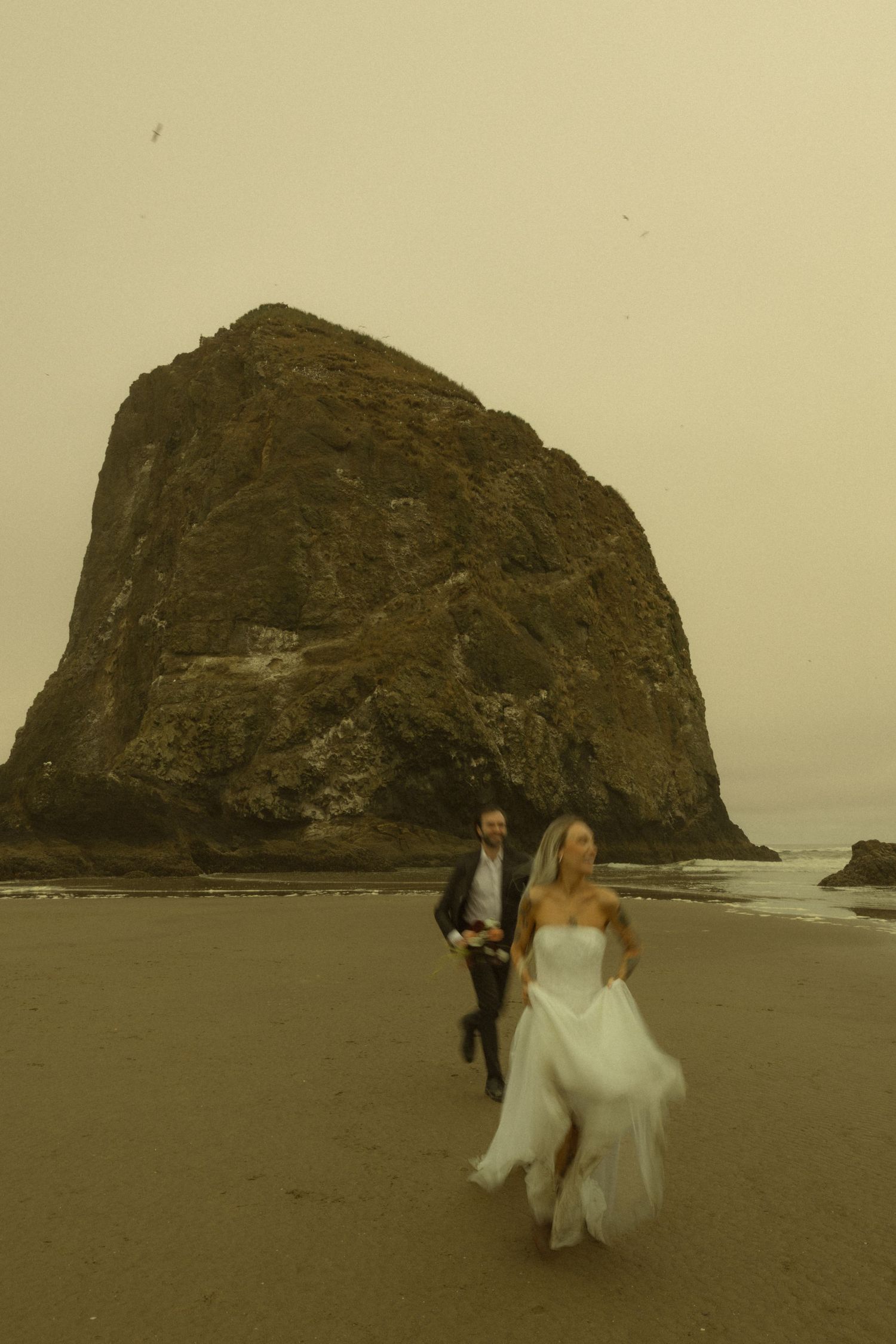 Bride and groom running on beach with Haystack Rock at Cannon Beach, Oregon on their wedding day.