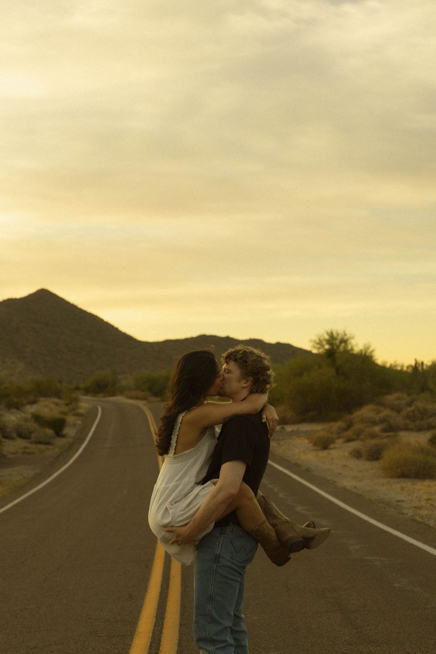 Romantic couple kissing on a desert road at golden hour with scenic mountain landscape.