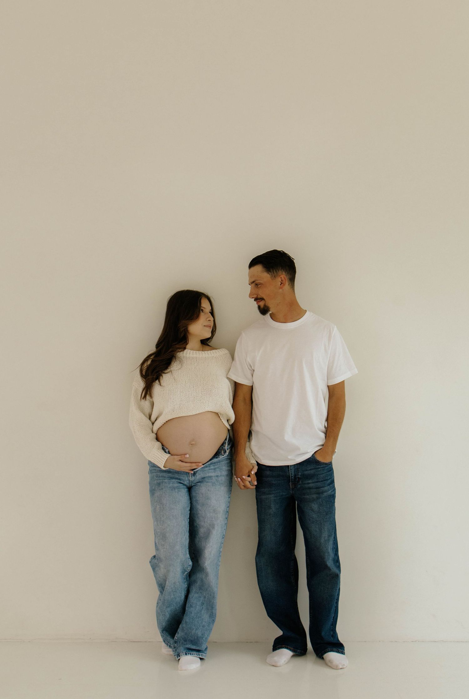 Expectant couple in casual denim and white tops stand against neutral wall in minimalist maternity photo.