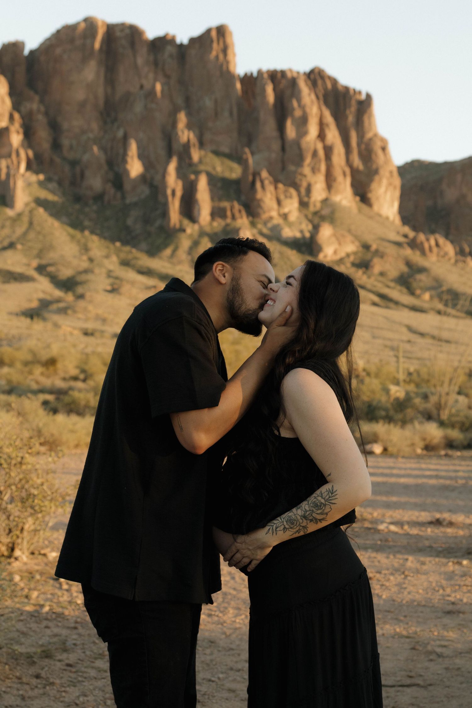 A romantic couple shares an intimate kiss in front of rugged desert mountains at sunset.