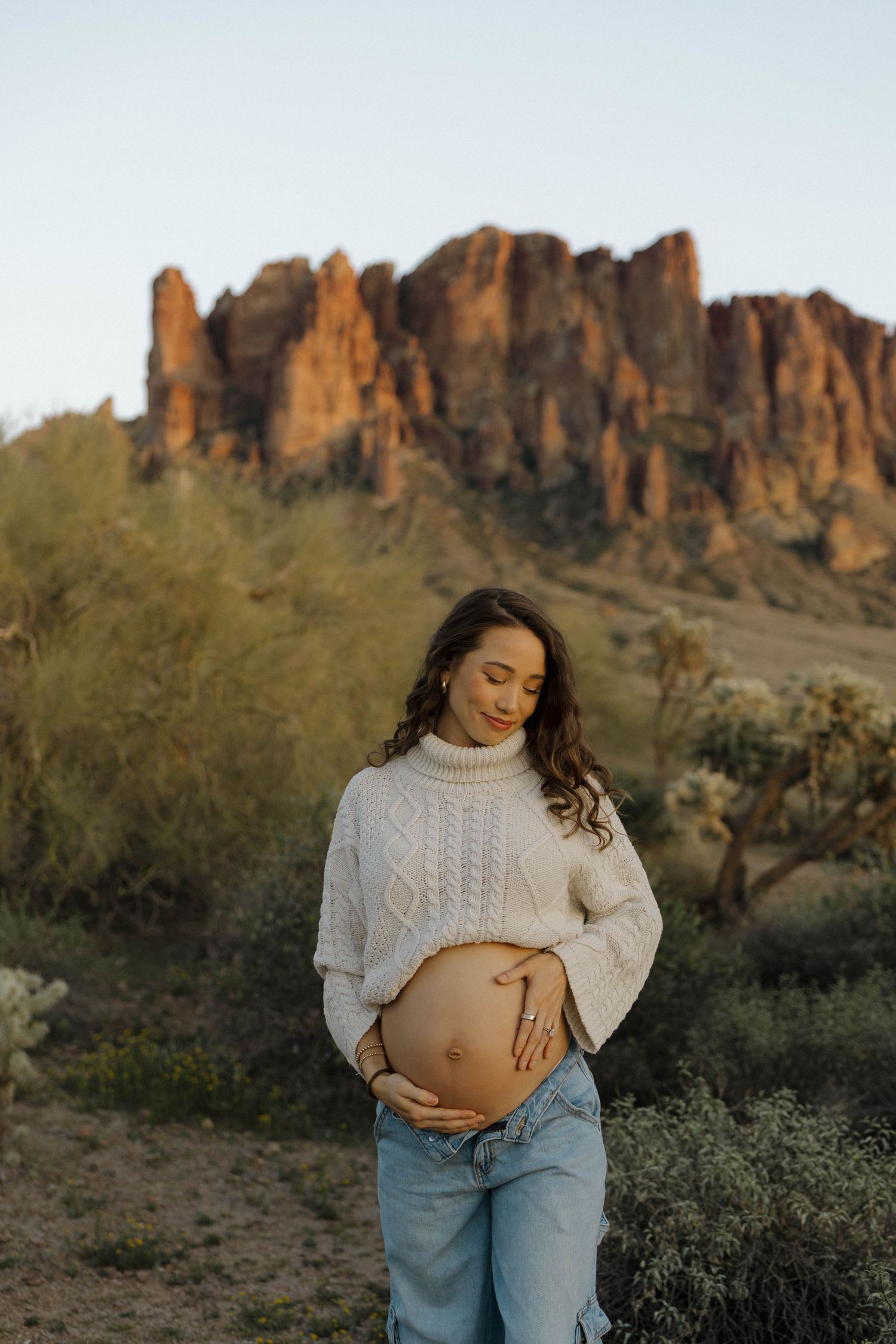 A person in a white sweater and jeans stands against a desert landscape with rocky mountains in the background.