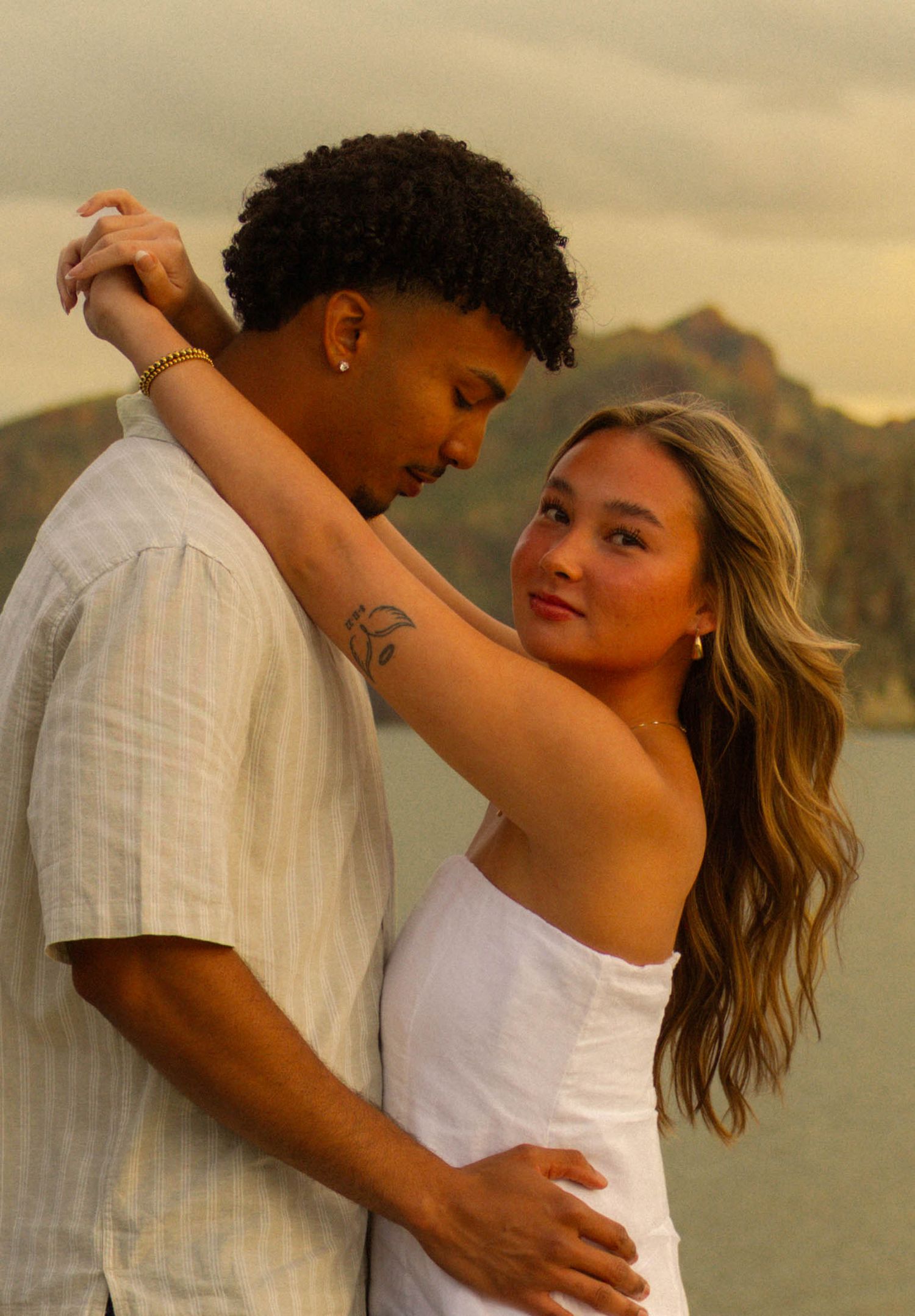 A romantic couple embraces at sunset with mountains in the background, dressed in white against a golden sky.