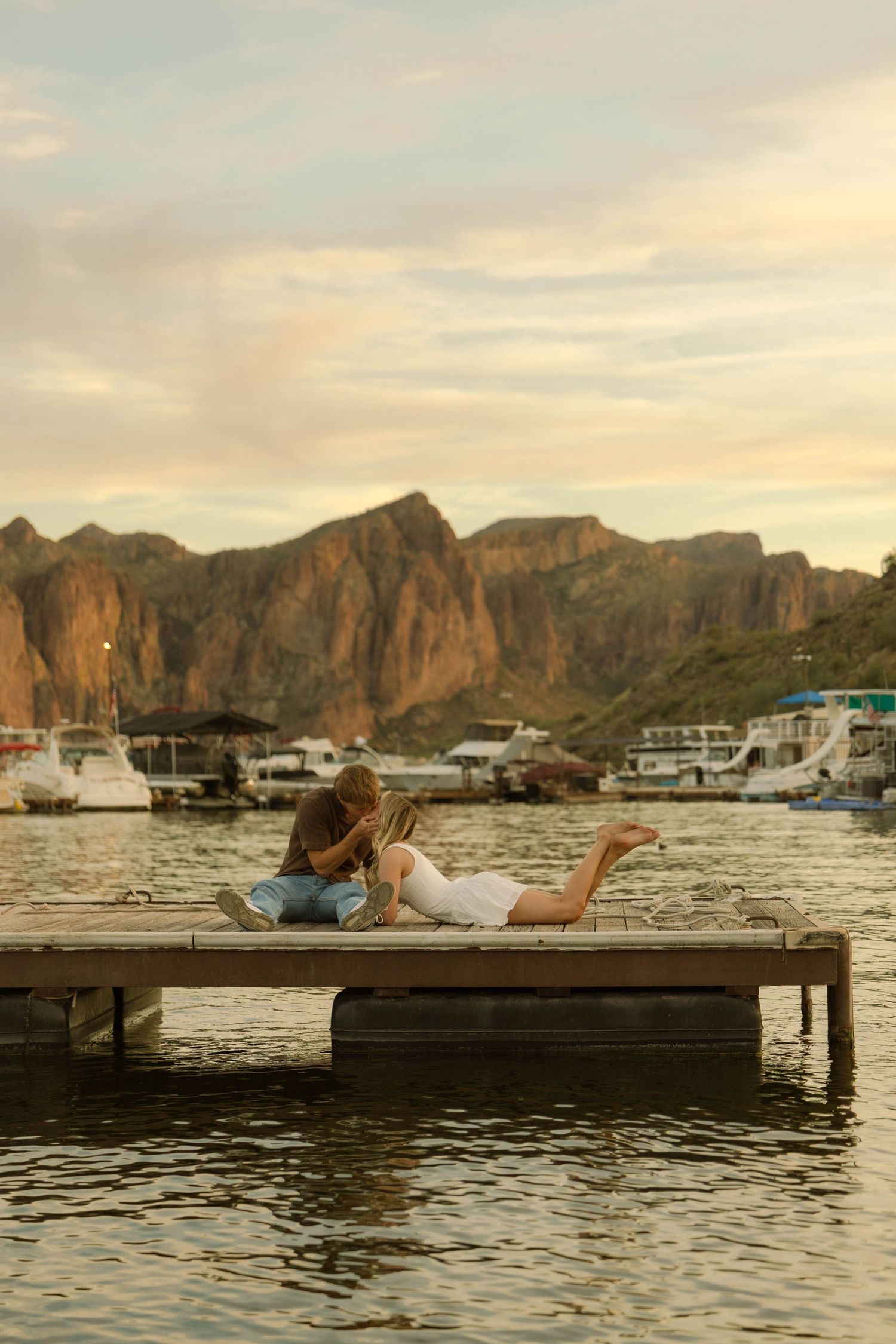 A couple lying on a wooden dock with dramatic mountain cliffs and boats in the background.