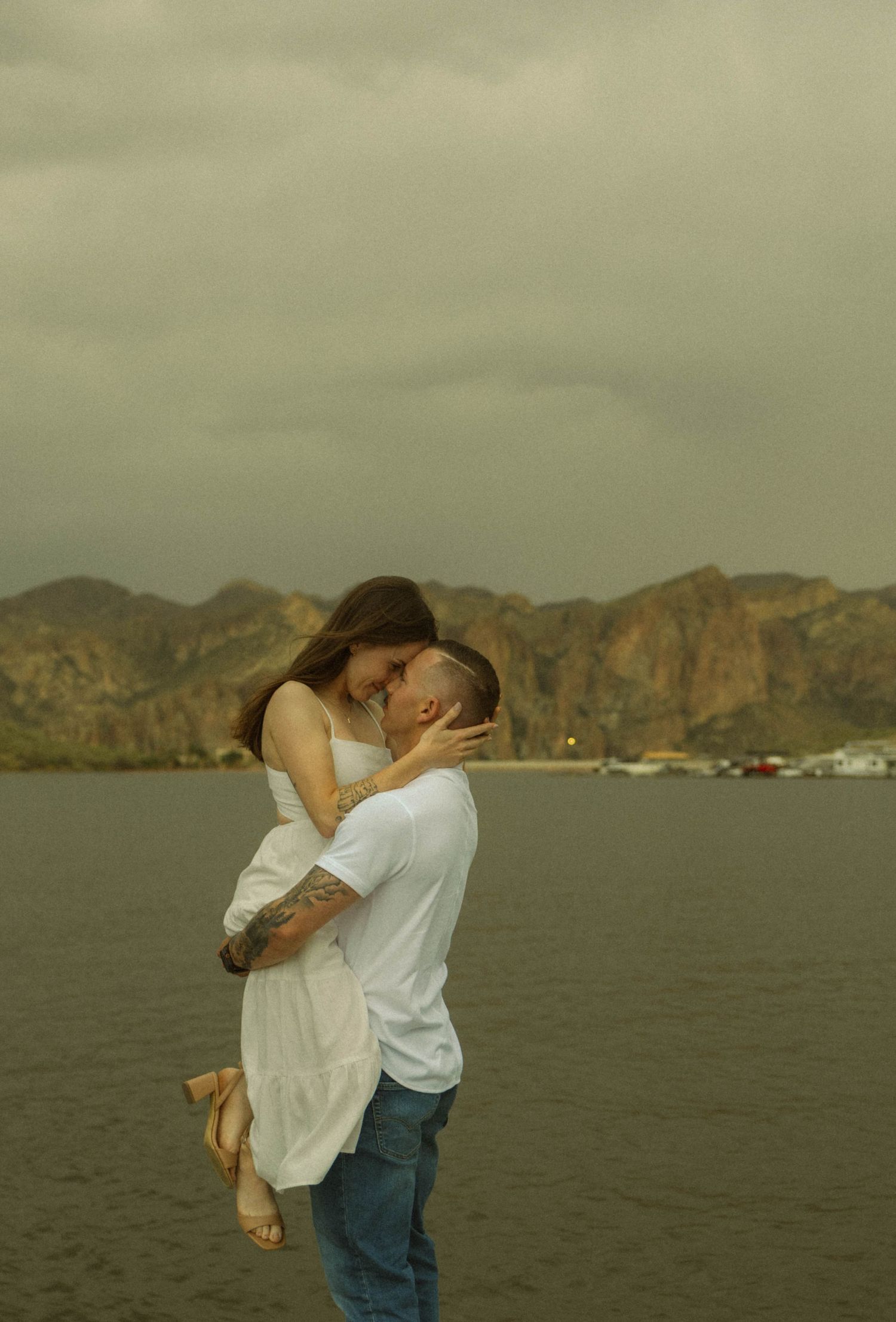 A couple embraces in a romantic moment against a scenic mountain and water backdrop at sunset.