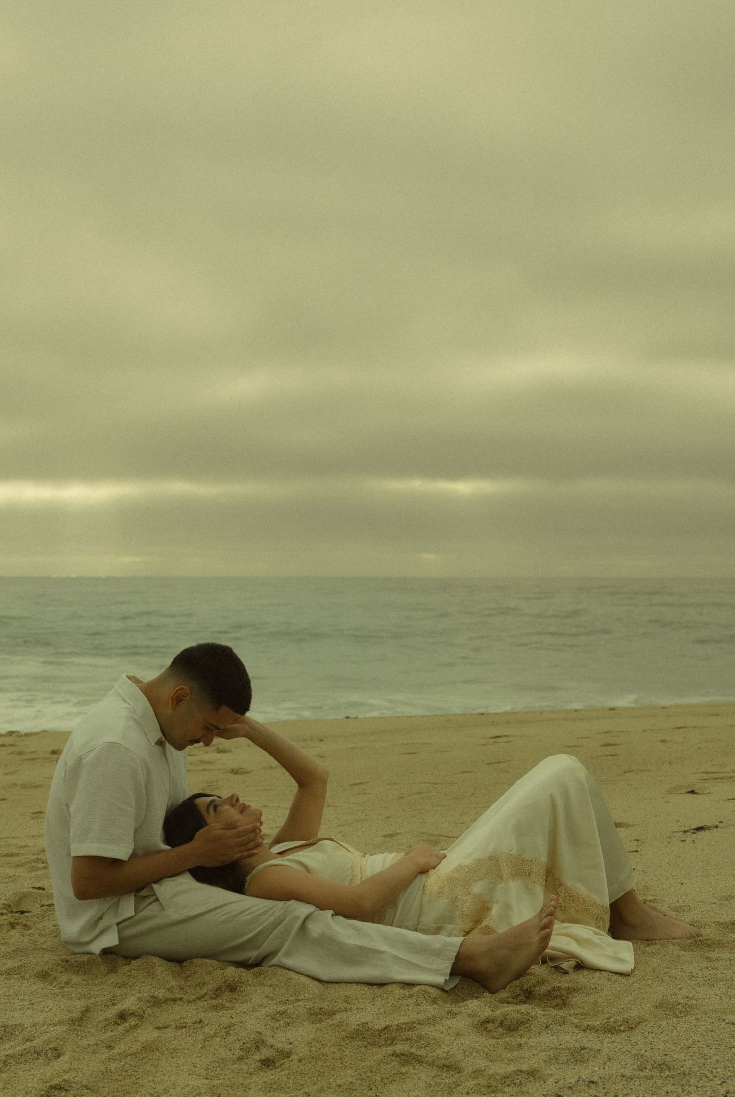 Couple sitting on sandy beach under overcast sky, woman reclining in white dress as man leans close romantically.
