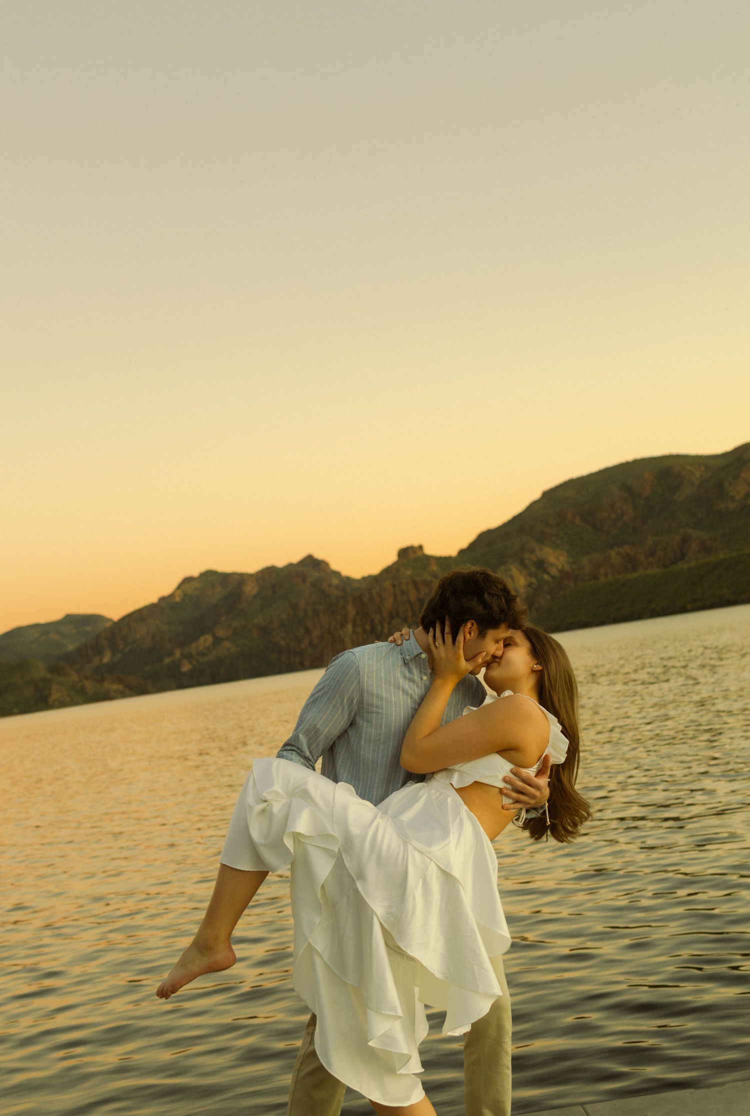 Romantic couple embracing at sunset by a serene lake with mountains in the background.