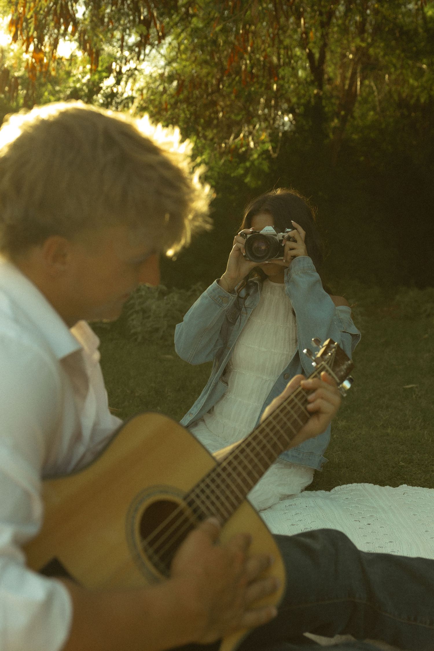 Woman with vintage camera photographing man playing acoustic guitar on grass during golden hour.