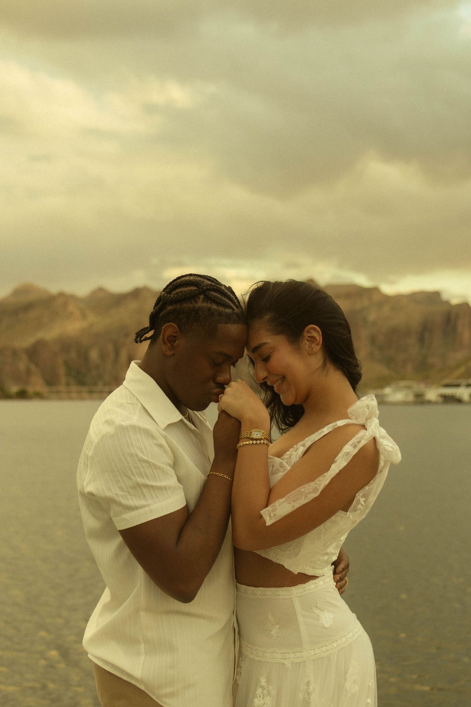 Interracial couple in white outfits sharing a romantic moment by a lake with mountains in the background.