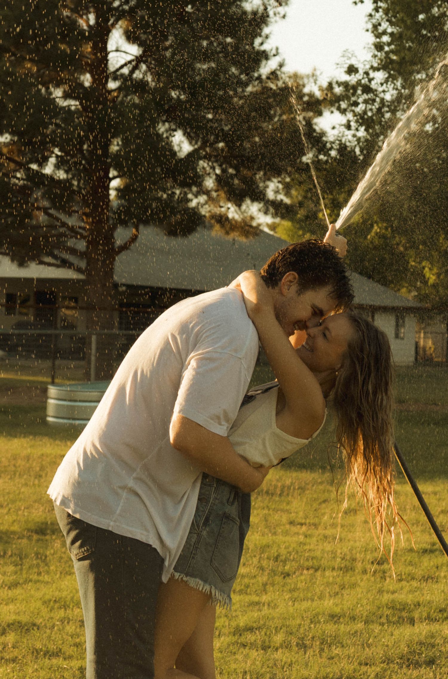 Couple kissing romantically in backyard while being sprayed by a garden hose on a warm sunny day.