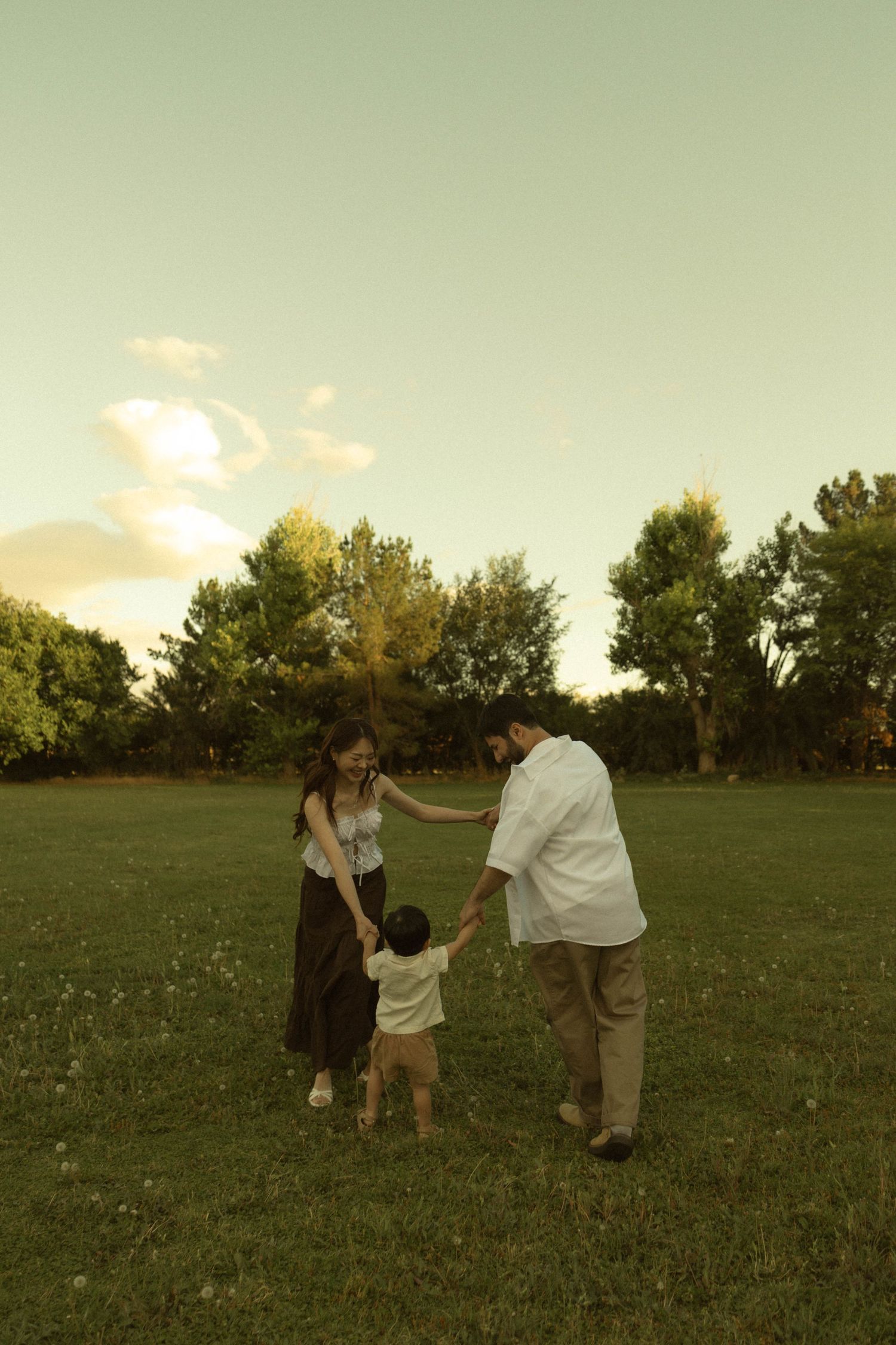 A young family holds hands with their toddler on a green lawn at sunset, surrounded by lush trees.