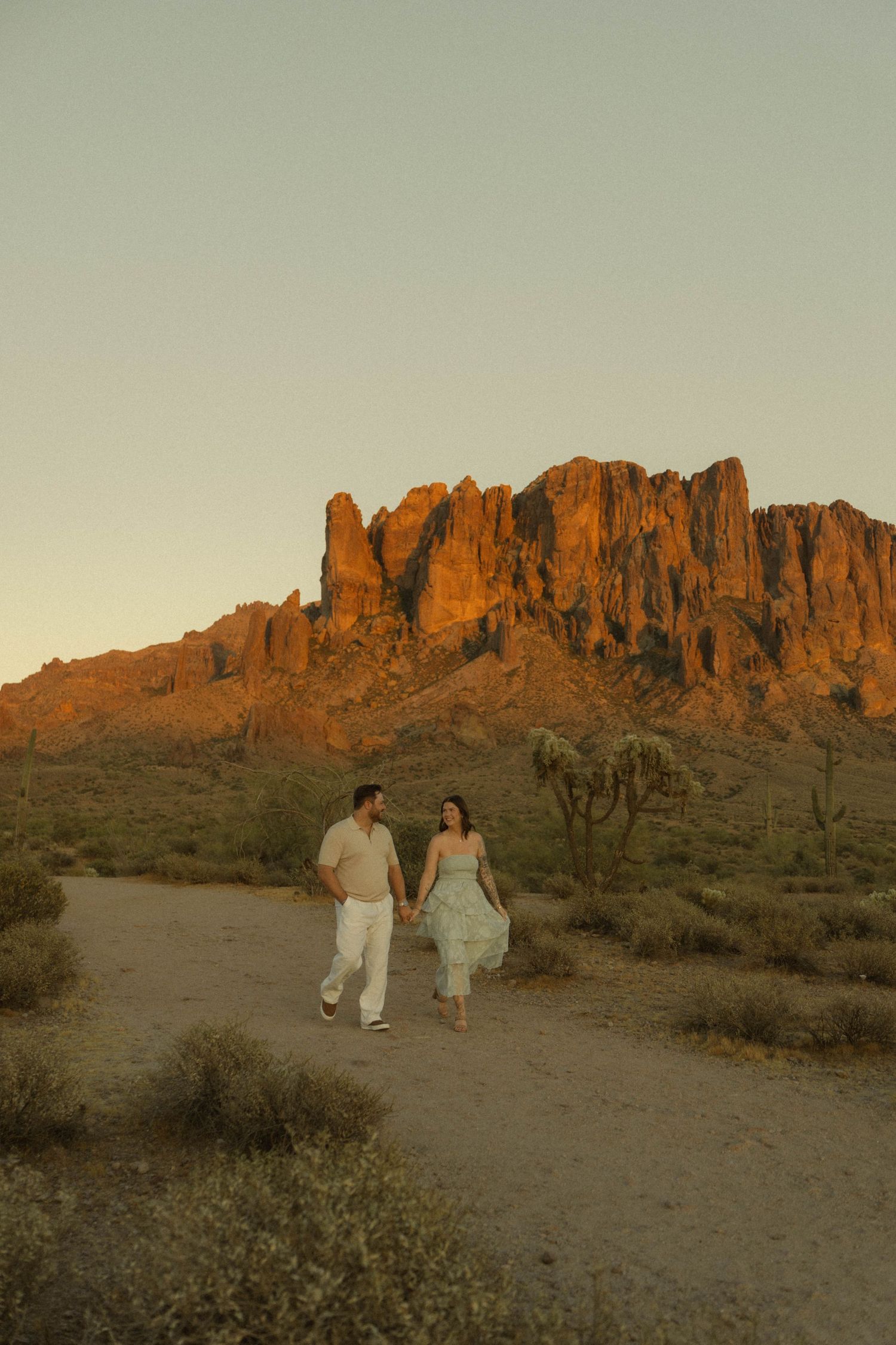 Couple holding hands walking in desert with dramatic Superstition Mountains glowing at sunset.