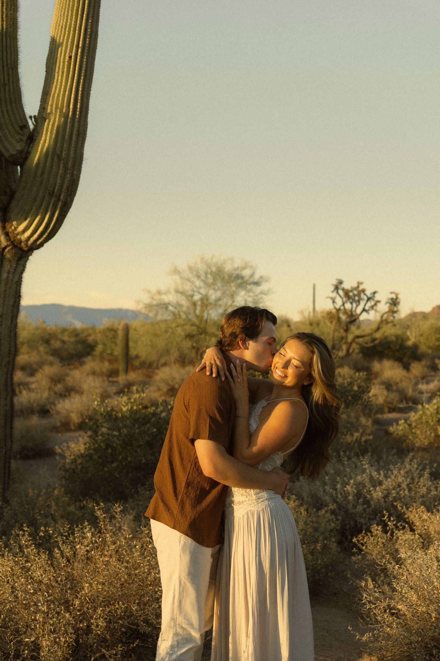 Couple embracing tenderly near a saguaro cactus in the warm golden light of the Arizona desert sunset.