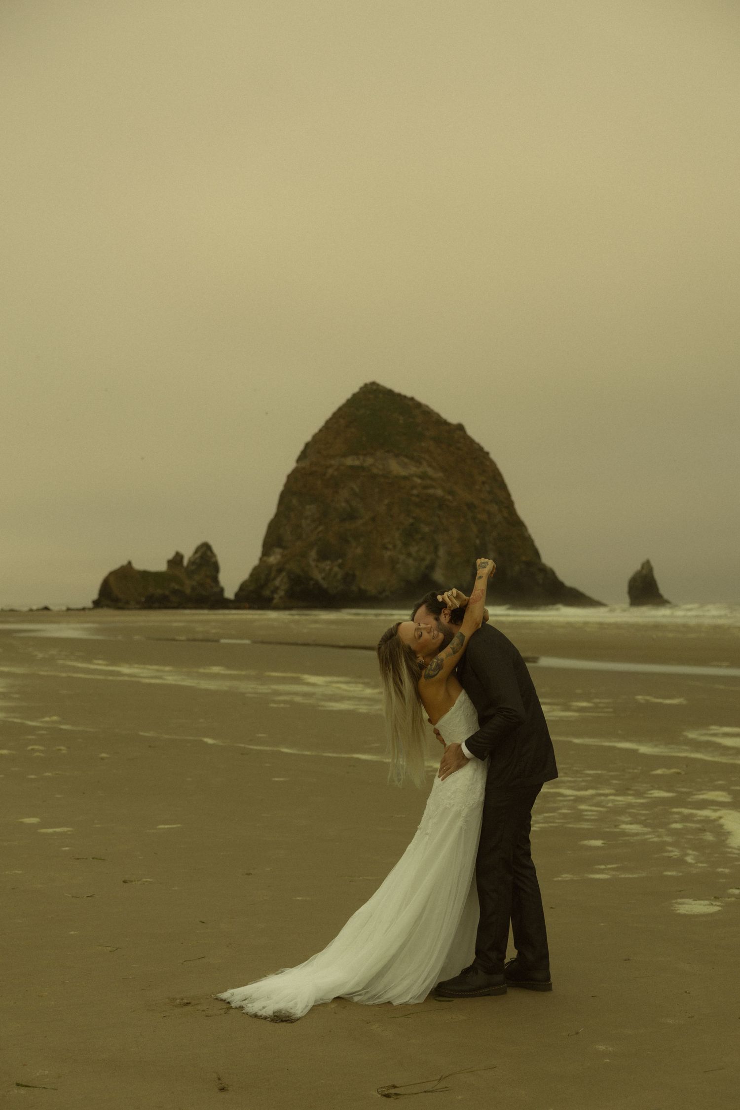 Bride and groom share a romantic kiss on a misty beach with Haystack Rock at Cannon Beach, Oregon in the background.