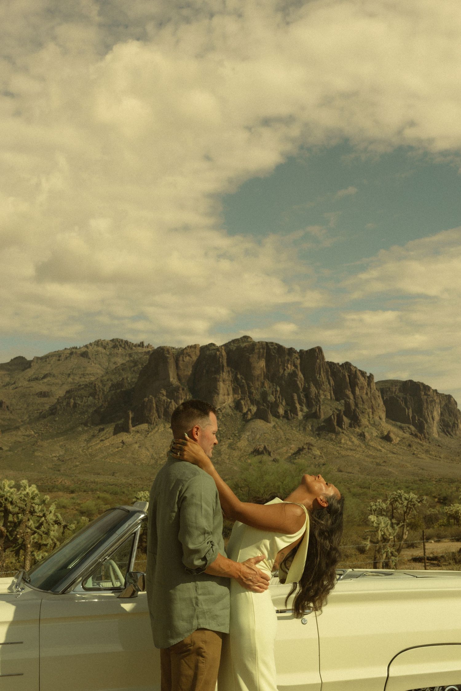 Couple embracing beside a vintage convertible car with Superstition Mountains and dramatic cloudy sky in Arizona desert.