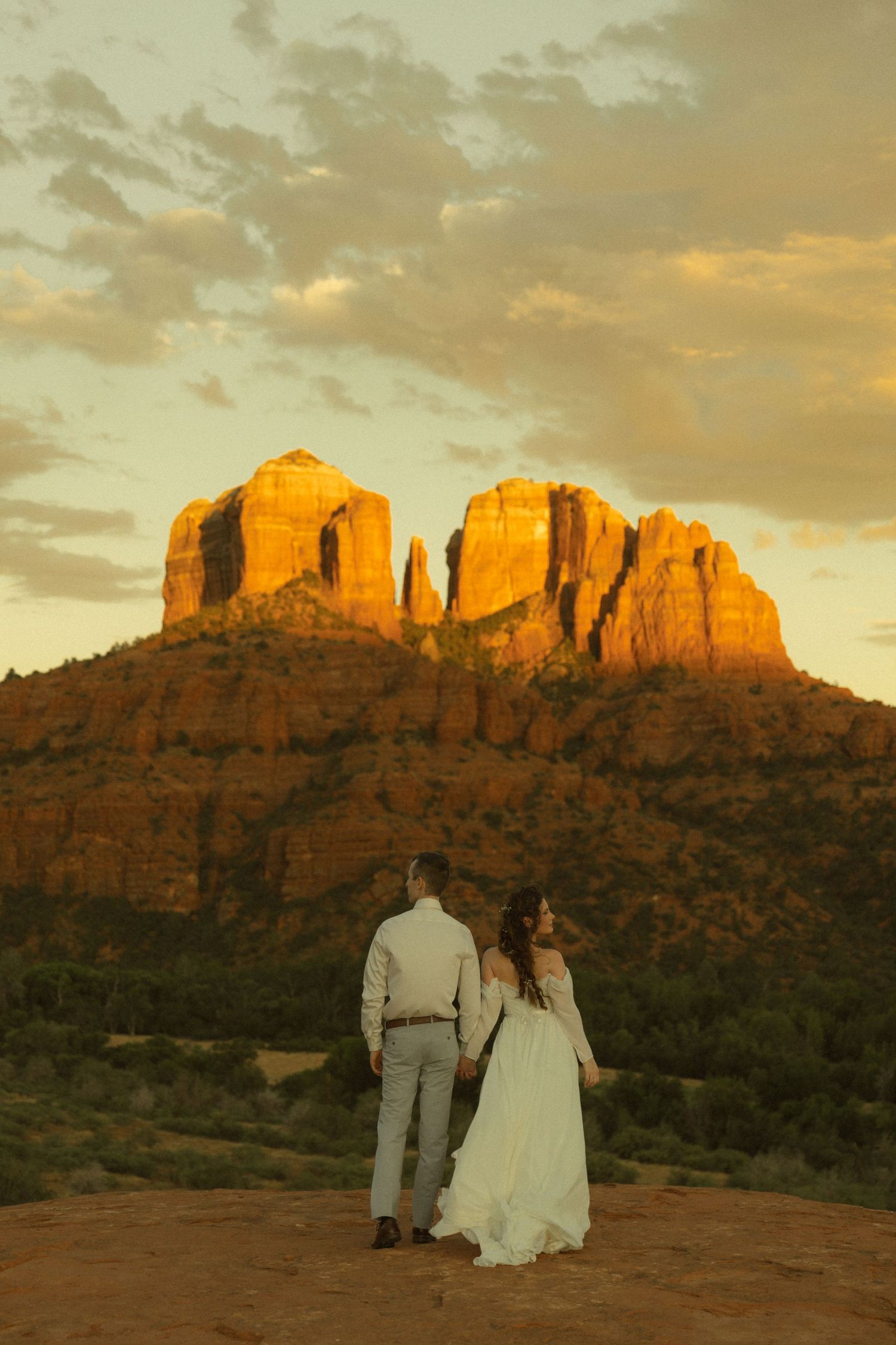 Couple in wedding attire holding hands facing Cathedral Rock at golden sunset in Sedona, Arizona.