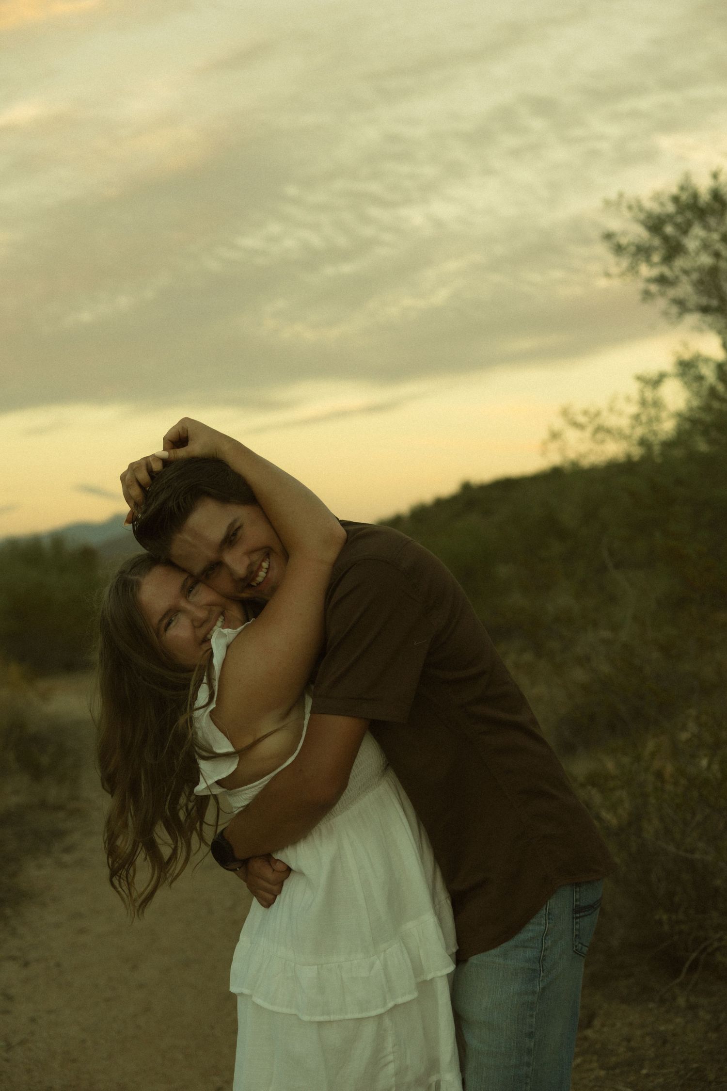 Couple embracing at sunset on a desert trail, woman in white dress laughing as man in brown shirt hugs her from behind.
