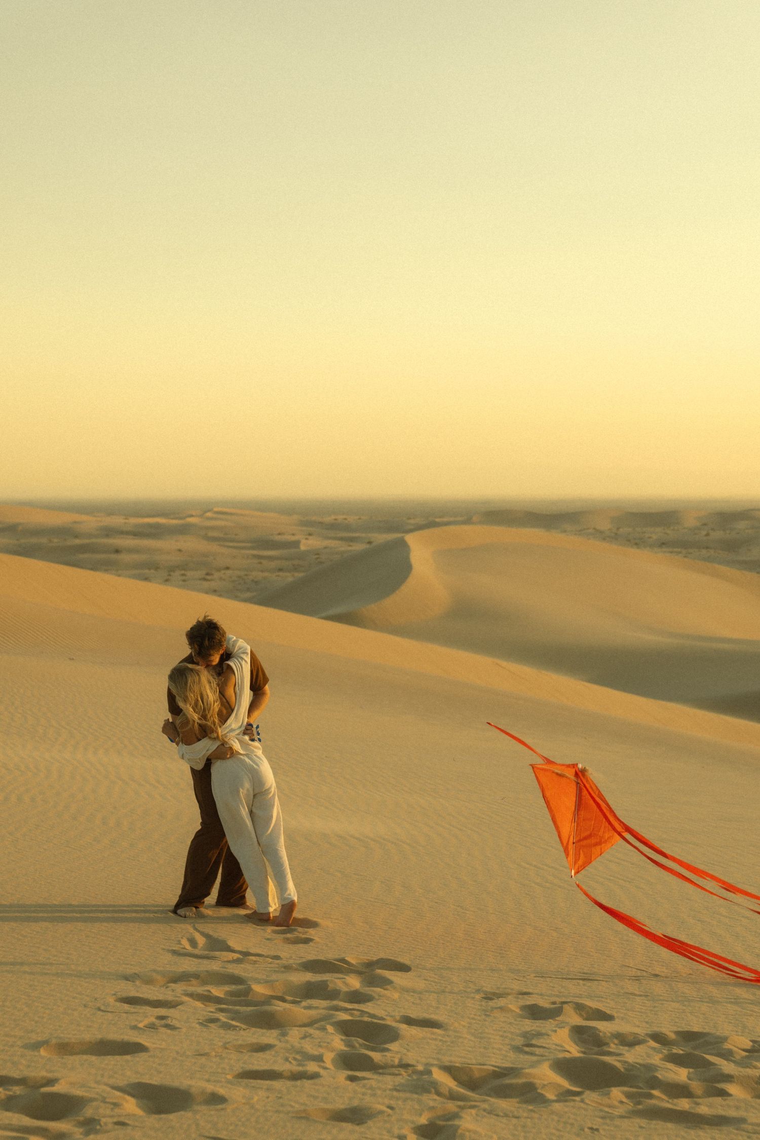 Couple embracing on golden sand dunes at sunset with a red kite flying nearby in the desert.