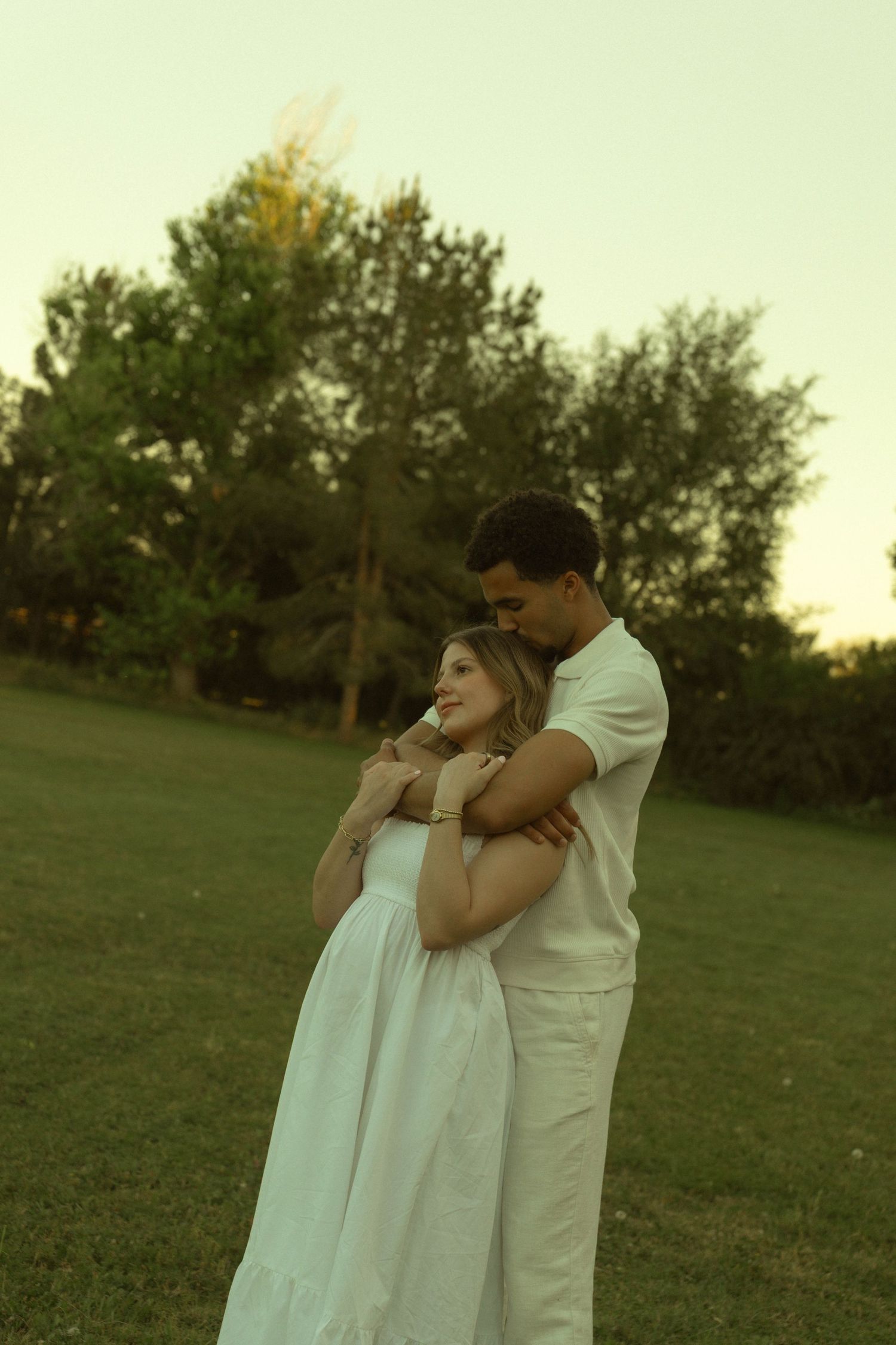 A couple embraces tenderly in a lush green park at sunset, wearing matching white outfits.