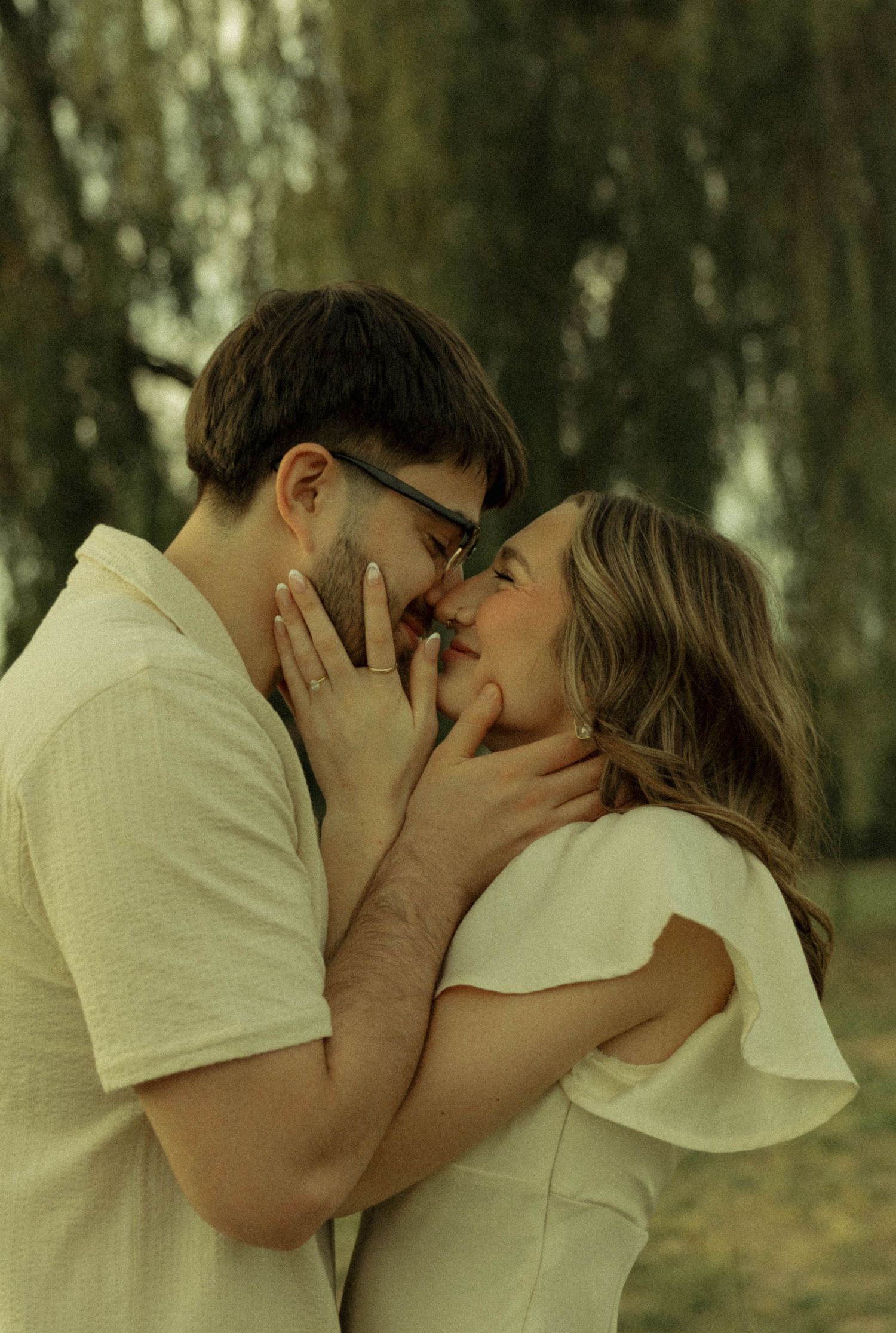 A couple shares a romantic almost-kiss outdoors, framed by soft green trees in a tender engagement photo.