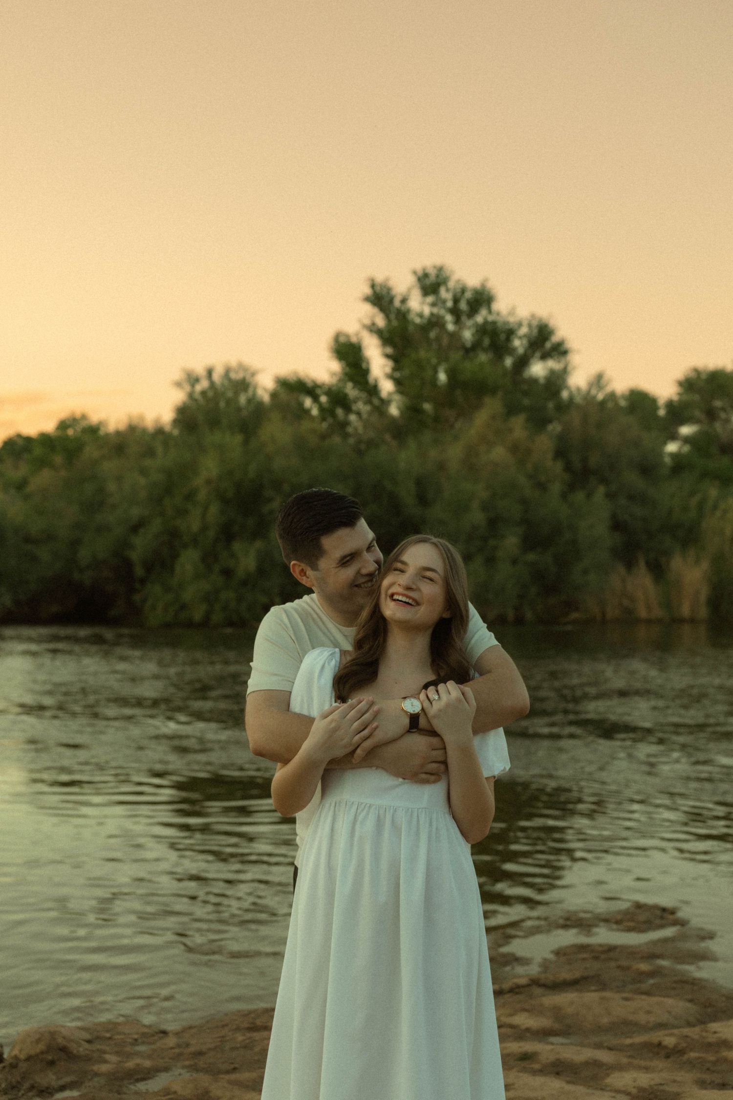 Happy couple embracing at golden hour sunset by a river, woman in white dress smiling joyfully.