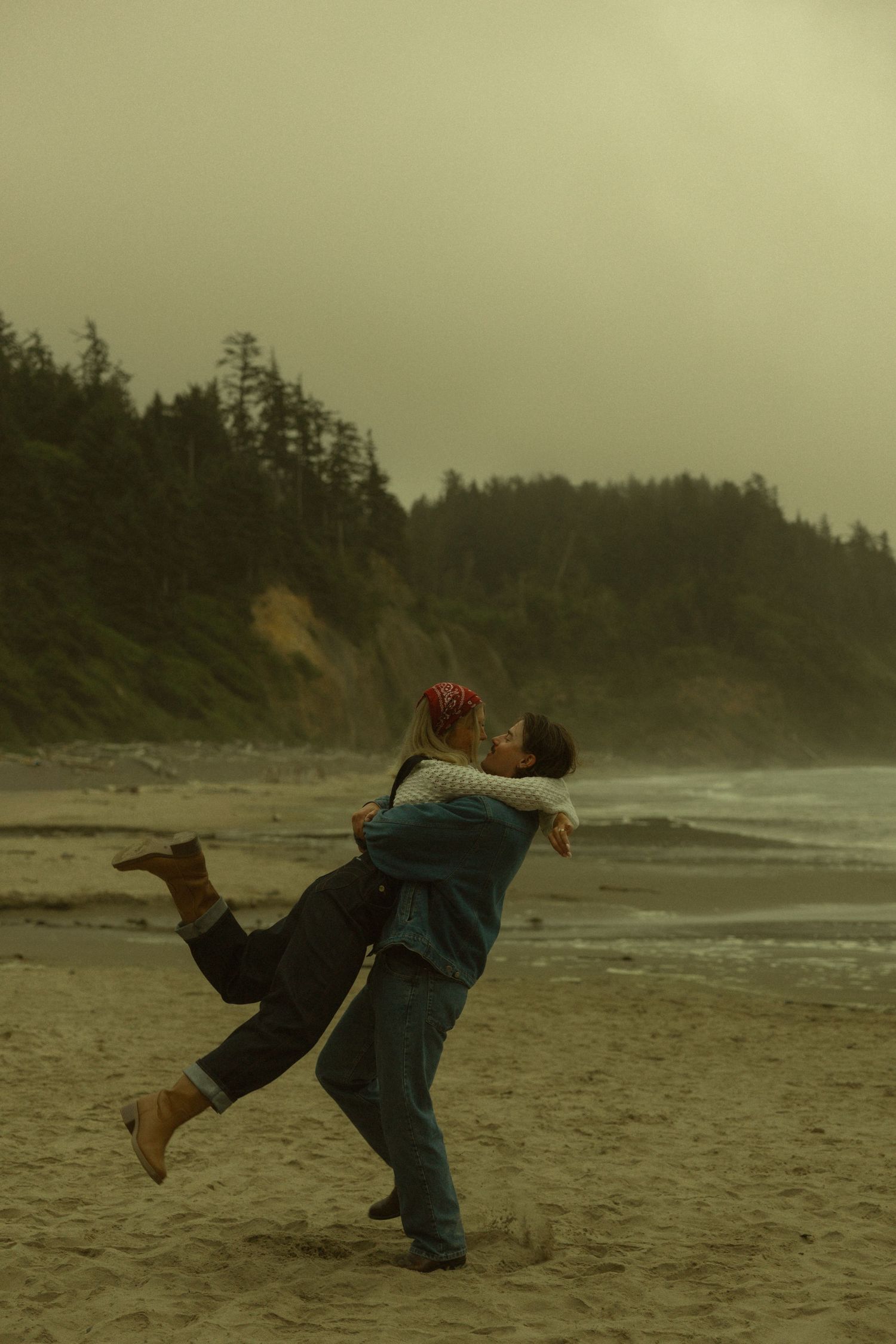 Couple embracing and spinning joyfully on a misty beach with evergreen cliffs and ocean waves in the background.