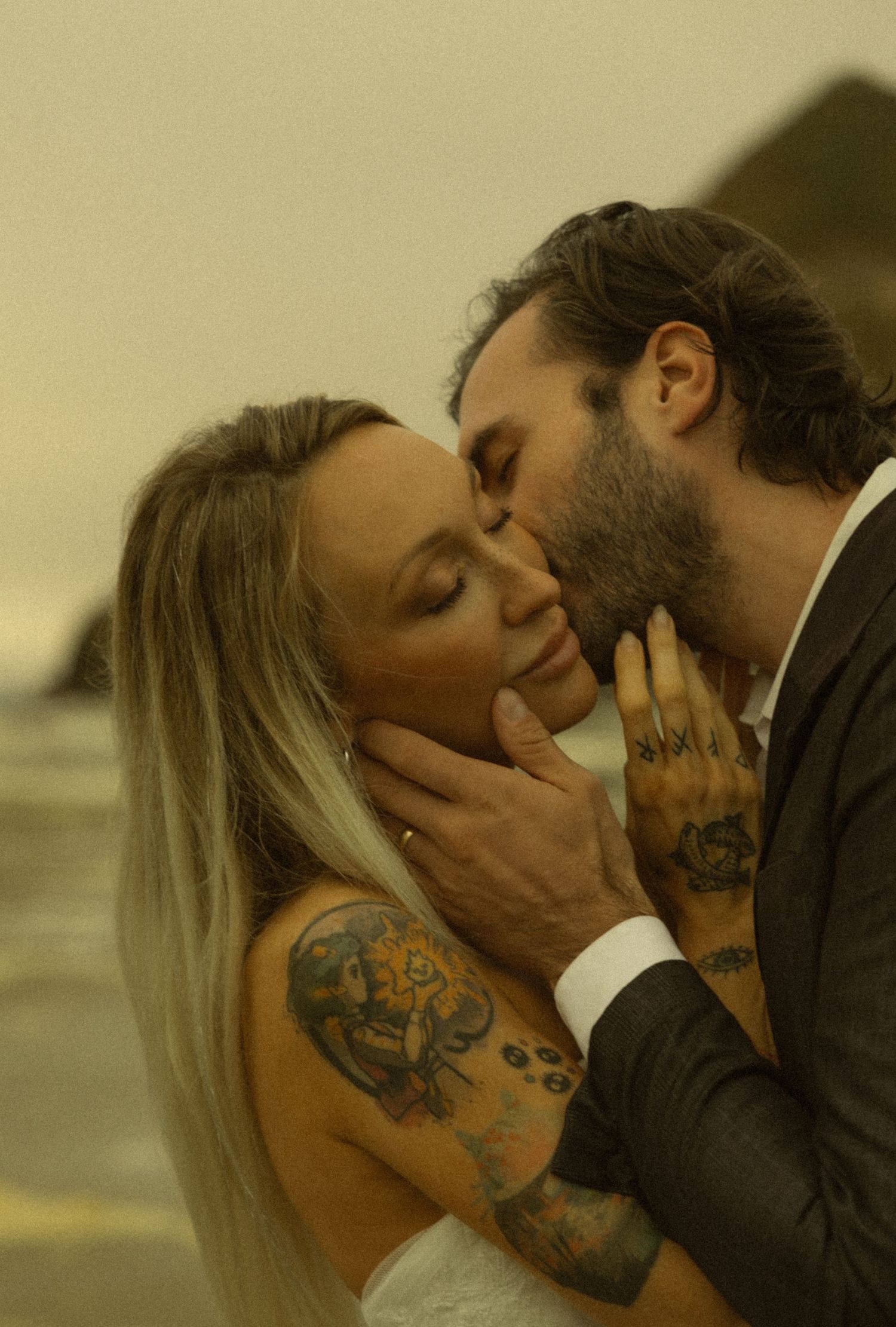 Groom tenderly kisses tattooed bride on cheek during romantic beach wedding ceremony.