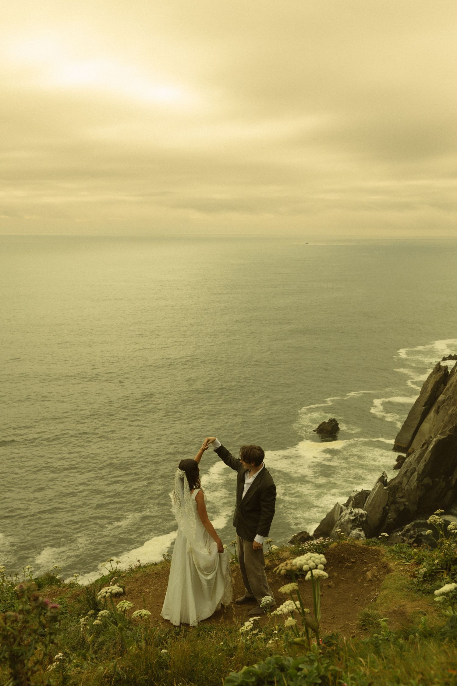 Bride and groom dancing on a cliff edge overlooking the ocean at sunset during their wedding photo shoot.