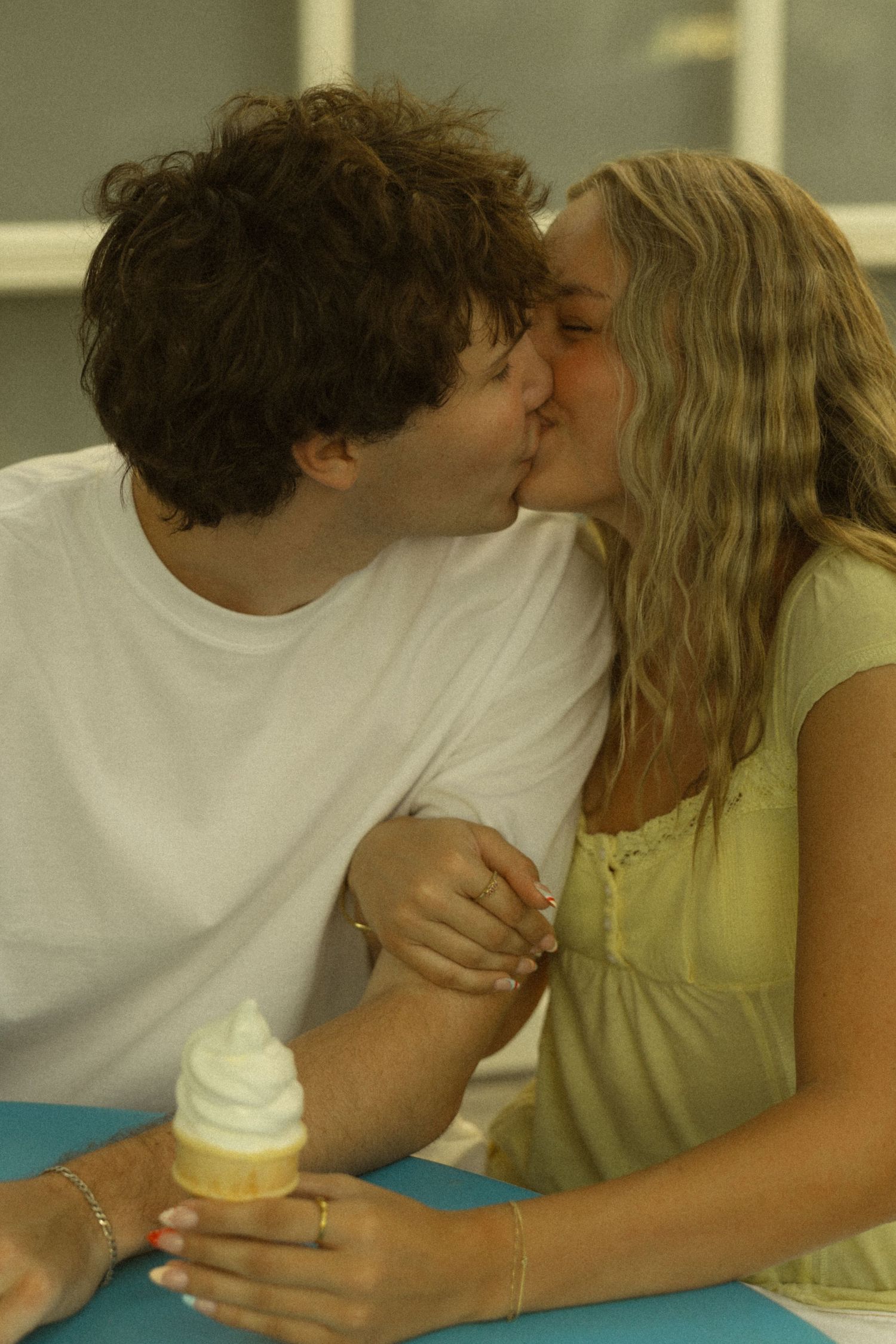 A young couple shares a romantic kiss while holding a soft serve ice cream cone at a casual dining table.