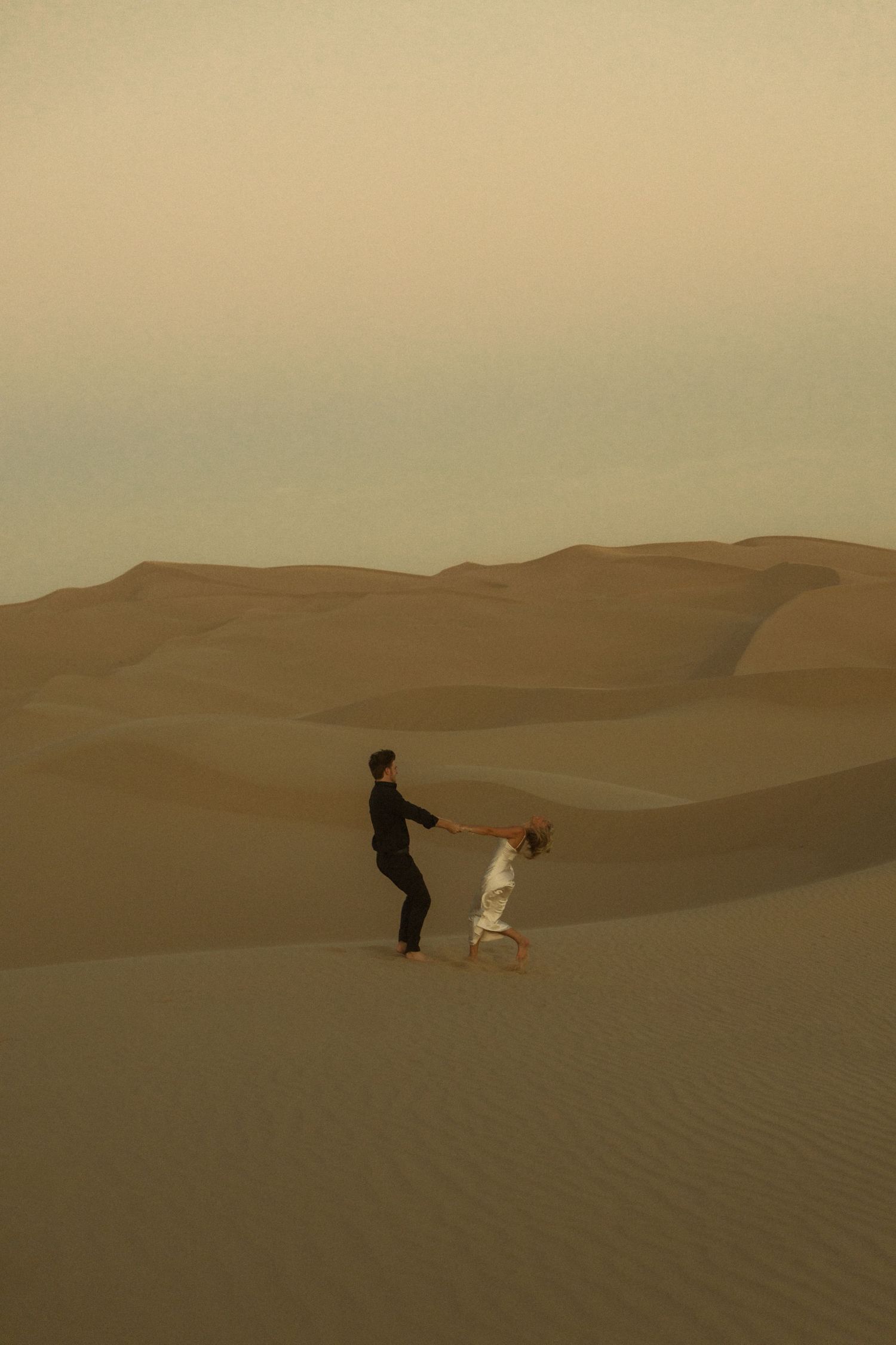 Couple playfully spinning on sandy desert dunes in hazy golden light.