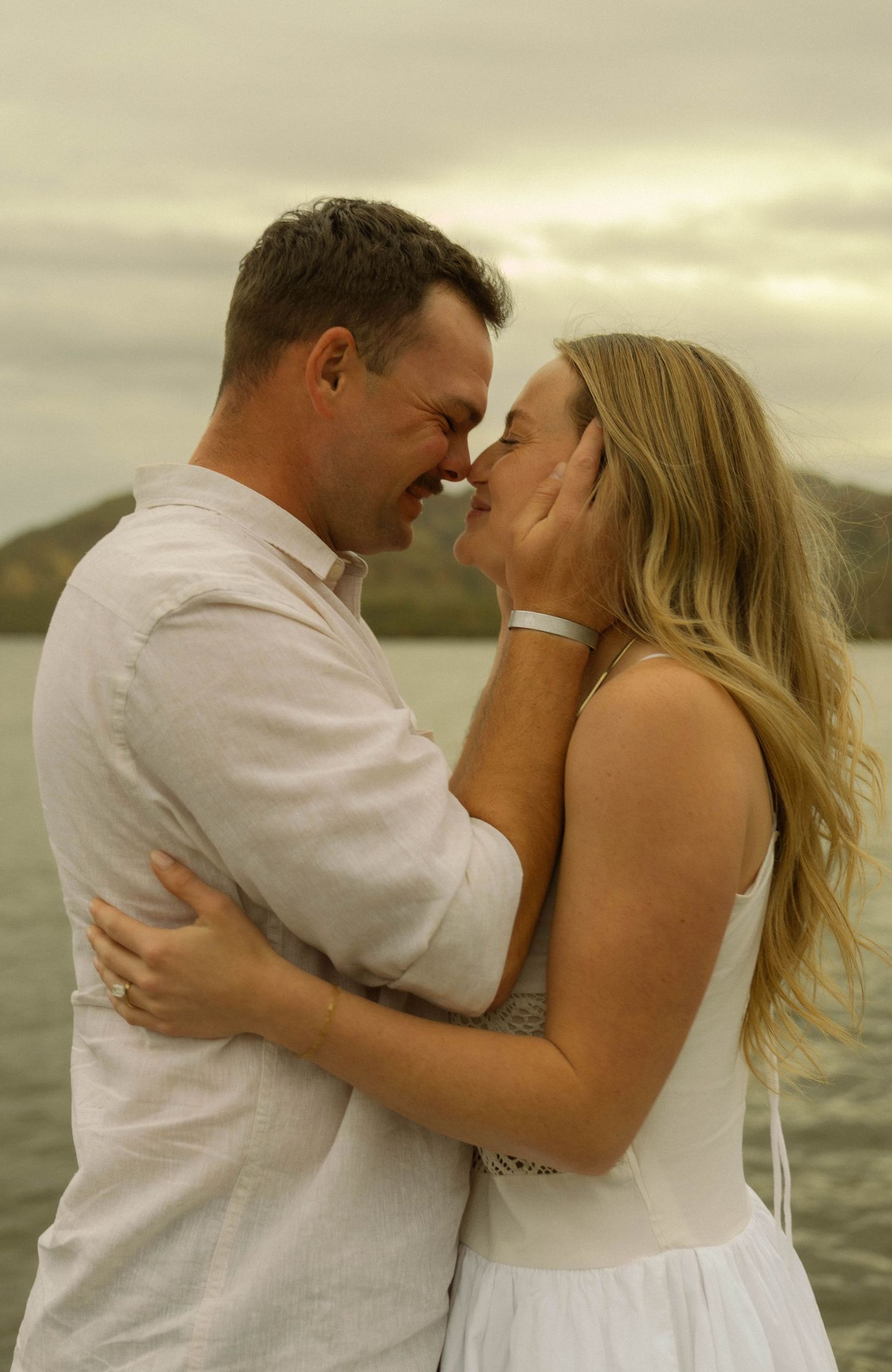 A tender embrace between a couple in white outfits against a cloudy coastal backdrop.