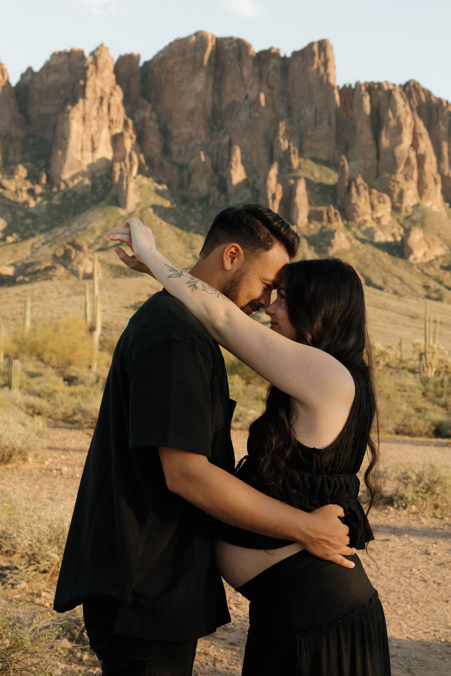A couple in dark clothing embraces against a backdrop of dramatic desert rock formations at sunset.
