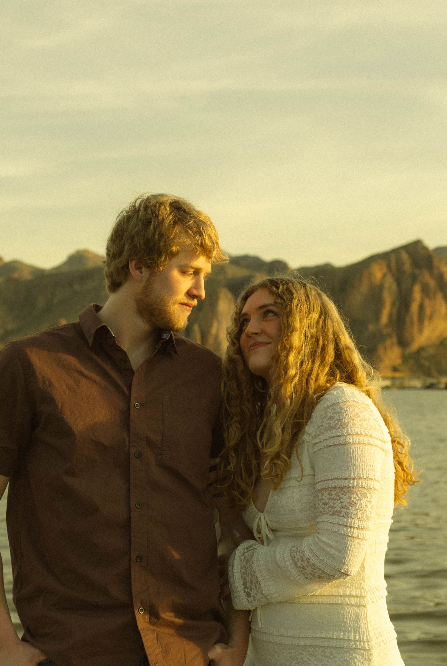 A couple shares an intimate moment at sunset by a lake with mountains in the background.