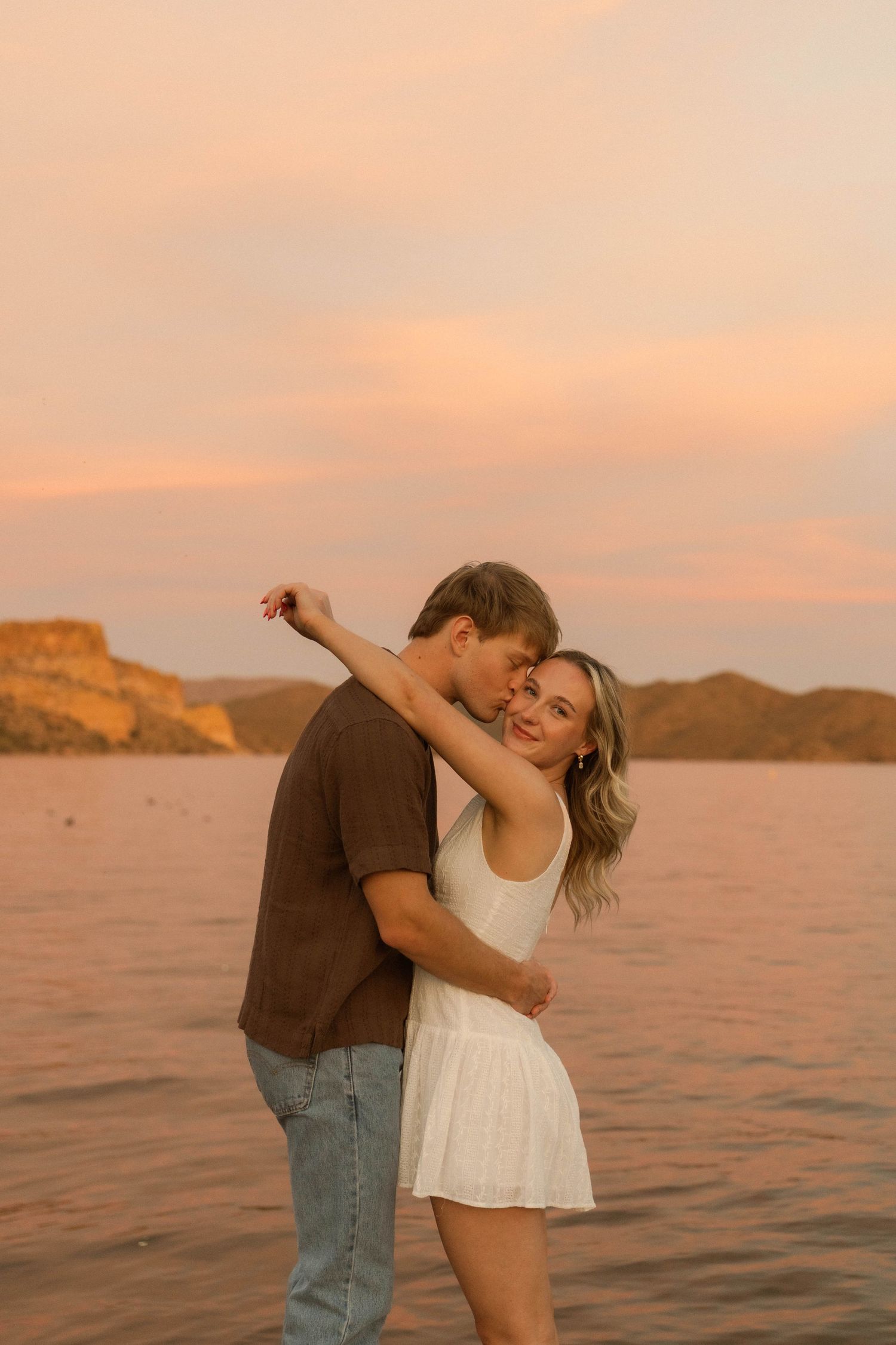 A couple shares a joyful dance moment on a dock with mountains and sunset waters behind them.