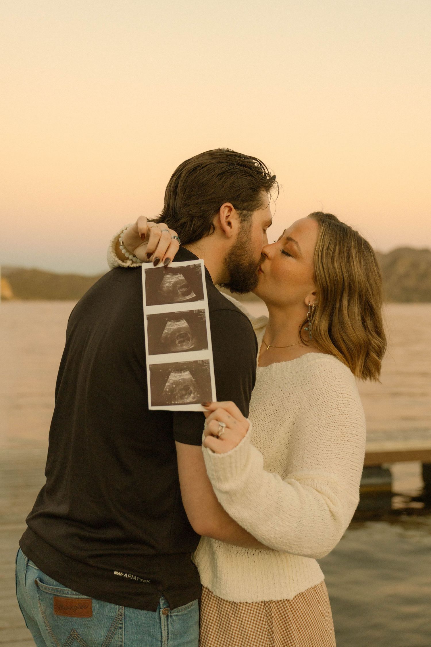 A couple shares a romantic kiss at sunset while holding an ultrasound photo near a waterfront dock.