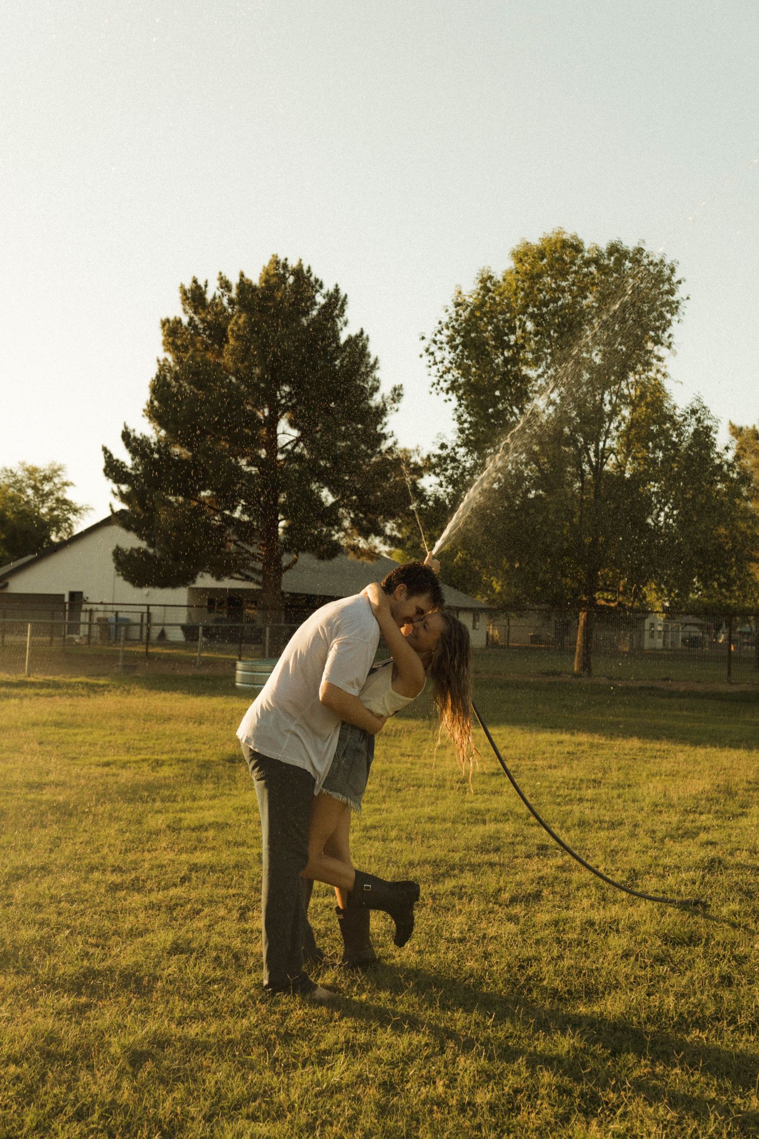 A couple shares a romantic kiss on a green lawn while a garden hose sprays water behind them.