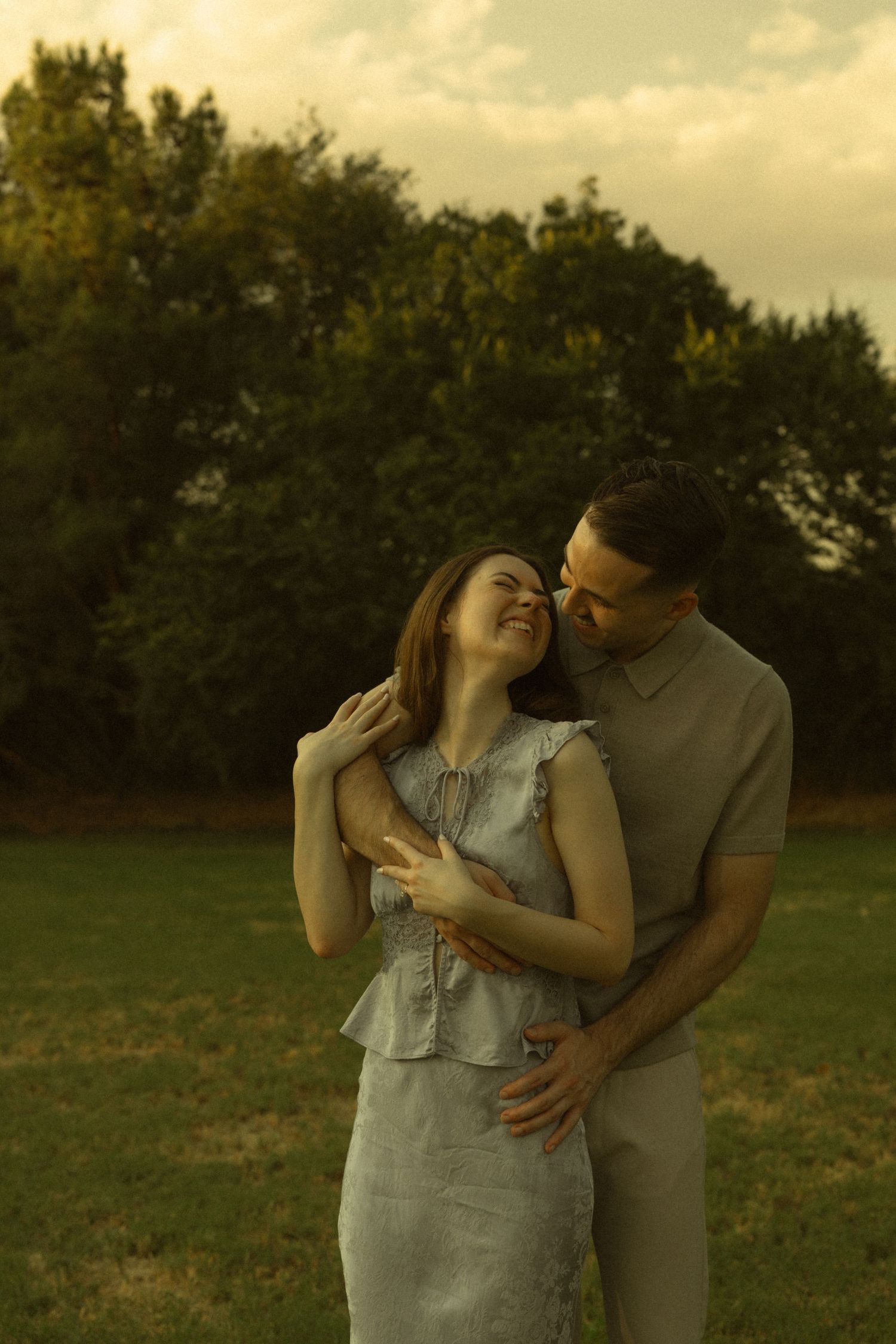Couple embracing in a park at golden hour, woman smiling as man kisses her cheek near lush green trees.