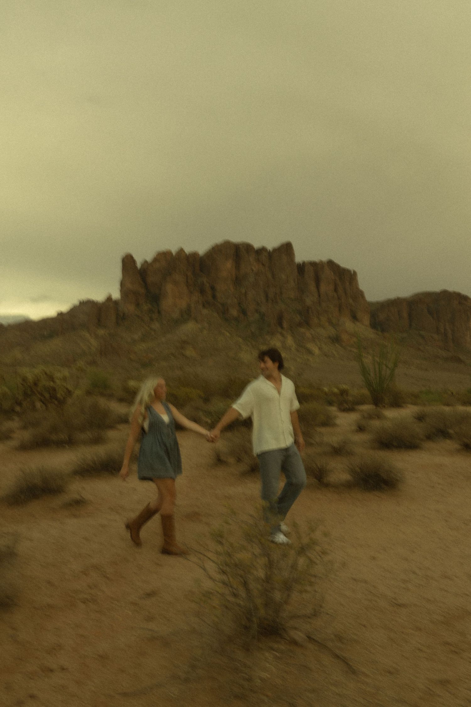 Couple holding hands and walking through the desert with rocky mountains under overcast skies.