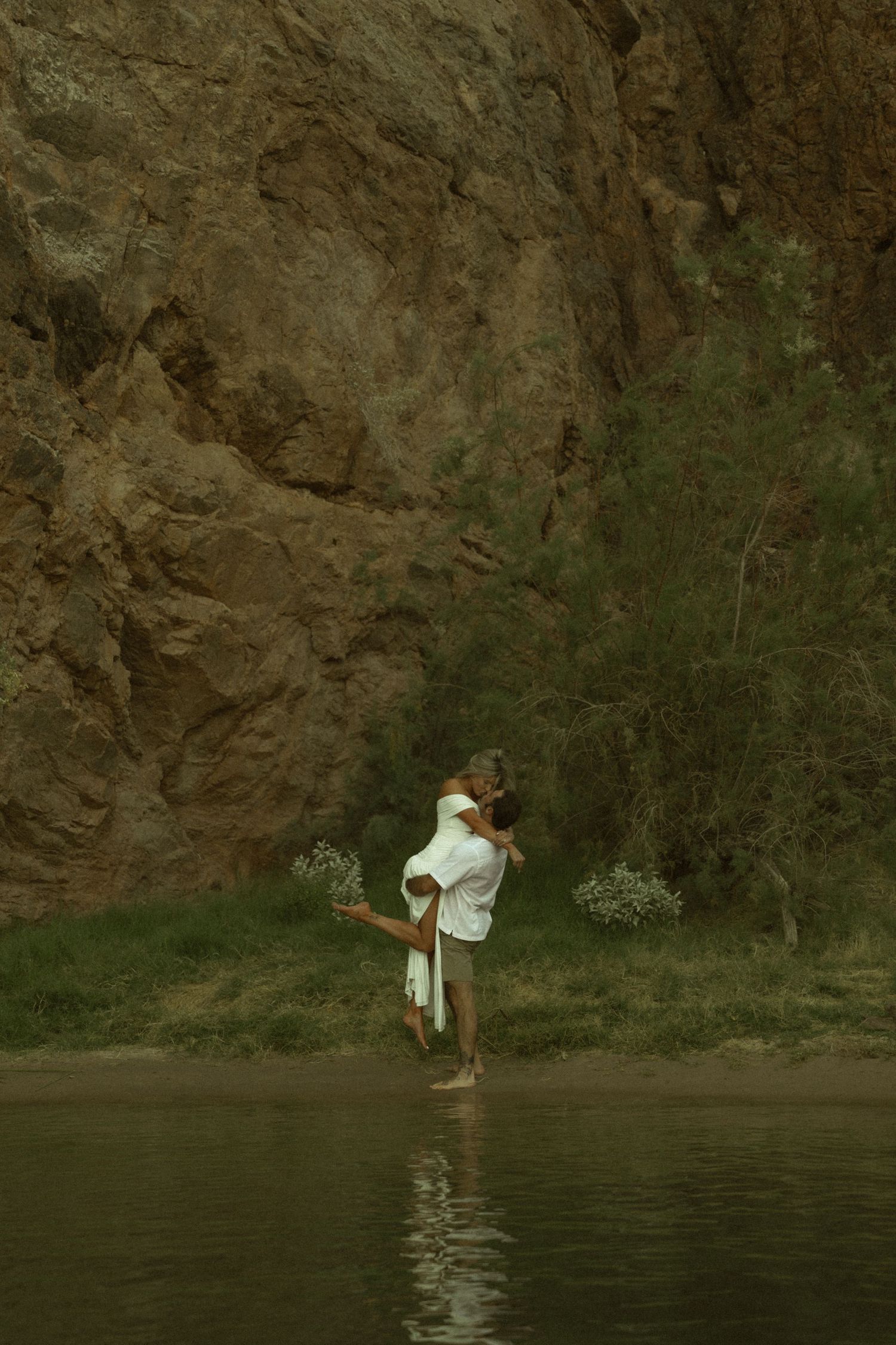 Romantic couple embracing by rocky canyon wall and lush greenery at tranquil river's edge.